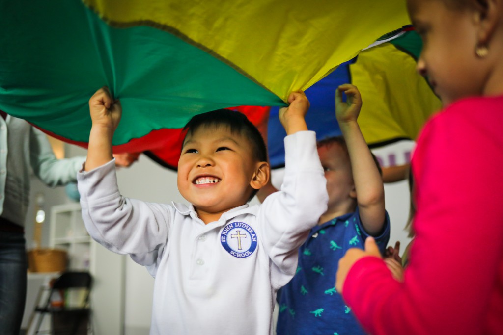 Child smiling and holding a colorful parachute with other children nearby indoors.