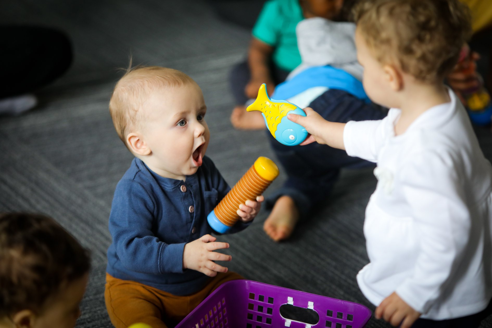 Two young children interact indoors, one holding a toy and the other reaching out with a fish-shaped toy, on a gray carpet.