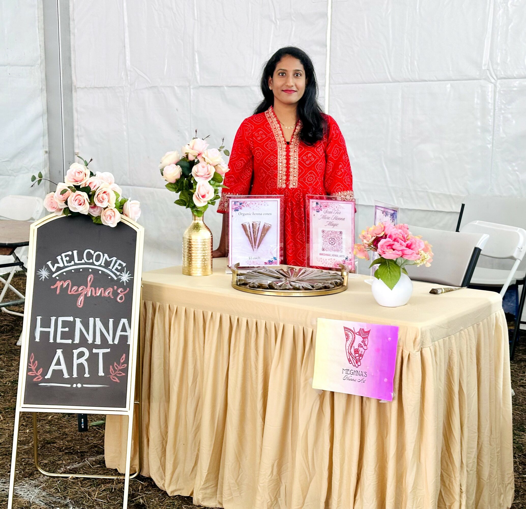 Woman in red traditional dress standing behind a table with henna art displays, flowers, and a welcome sign, inside a tent.