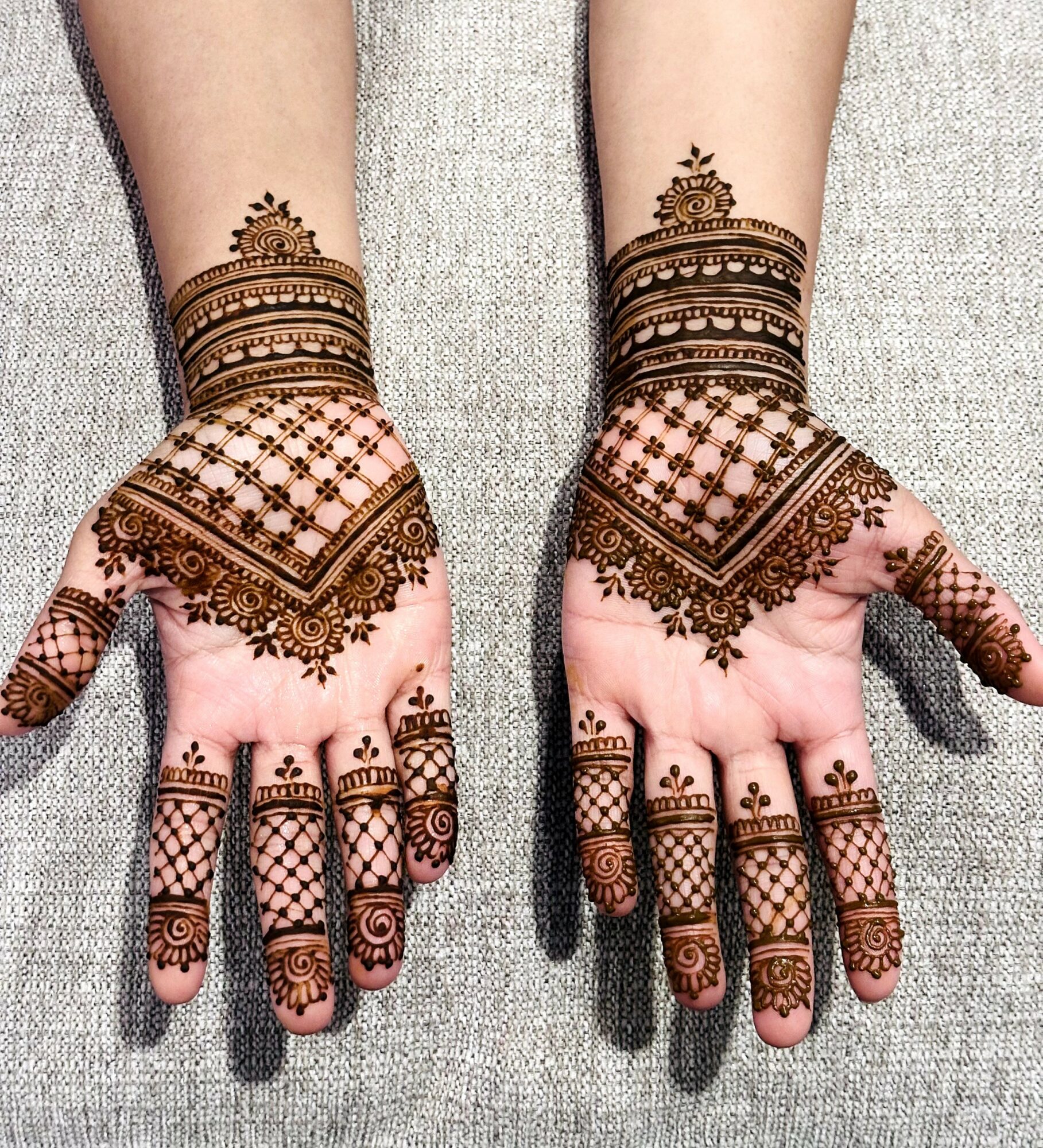Hands with intricate henna designs, resting on a gray surface, detailed patterns on fingers and wrists.