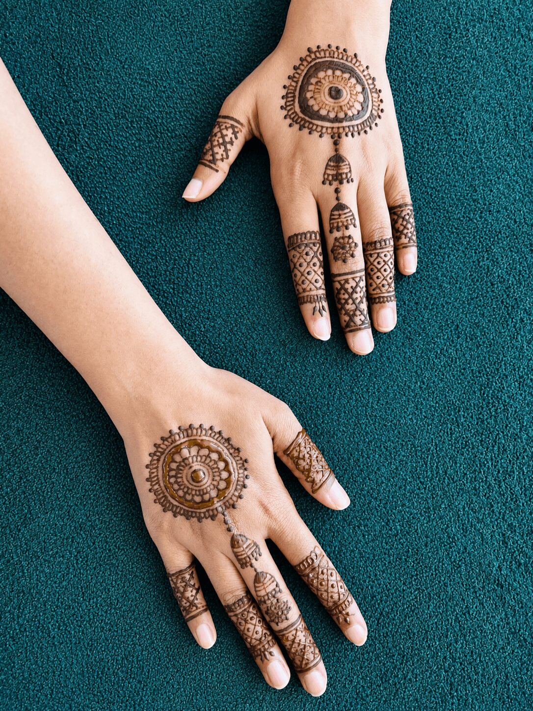 Two hands with intricate henna designs on fingers and backs, resting on a dark teal surface.