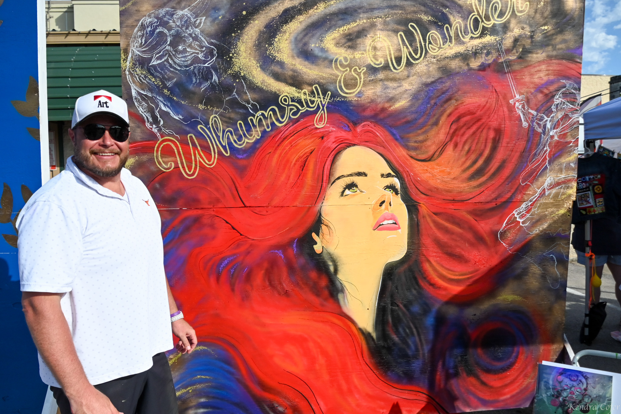 Man with sunglasses and white shirt standing next to a colorful mural of a woman with red hair and cosmic background.