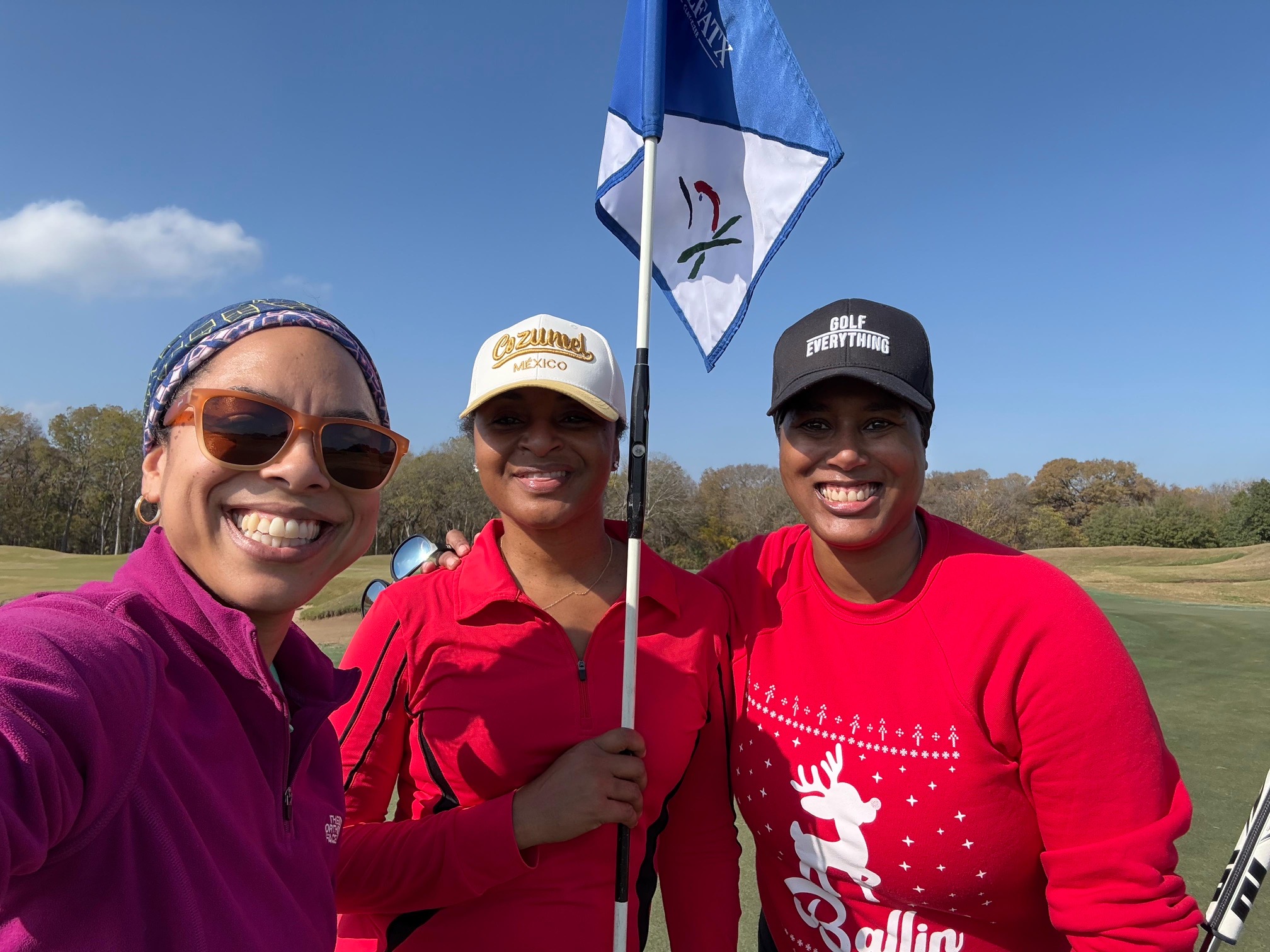 Three women smiling on a golf course, holding a flag, wearing hats and sunglasses, with a blue sky background.