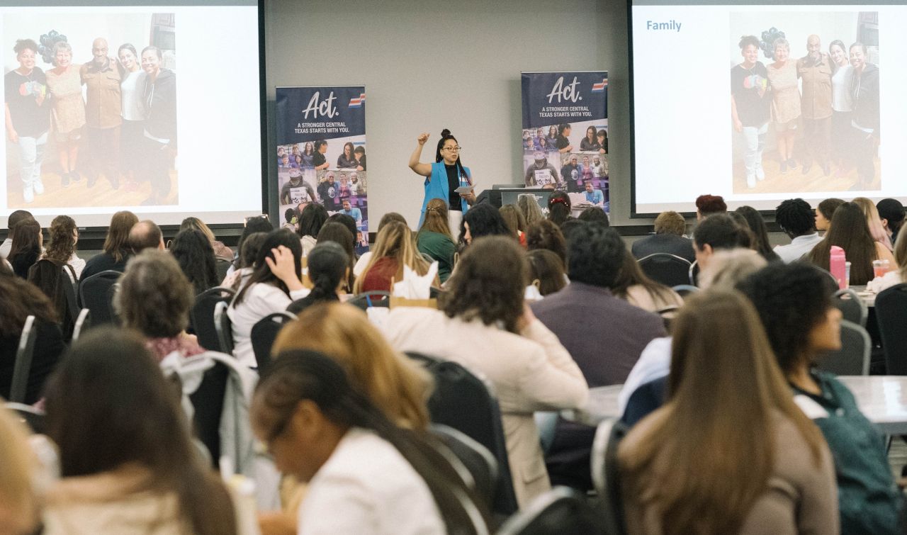 Speaker on stage presenting to a large audience in a conference room with two screens displaying a group photo.