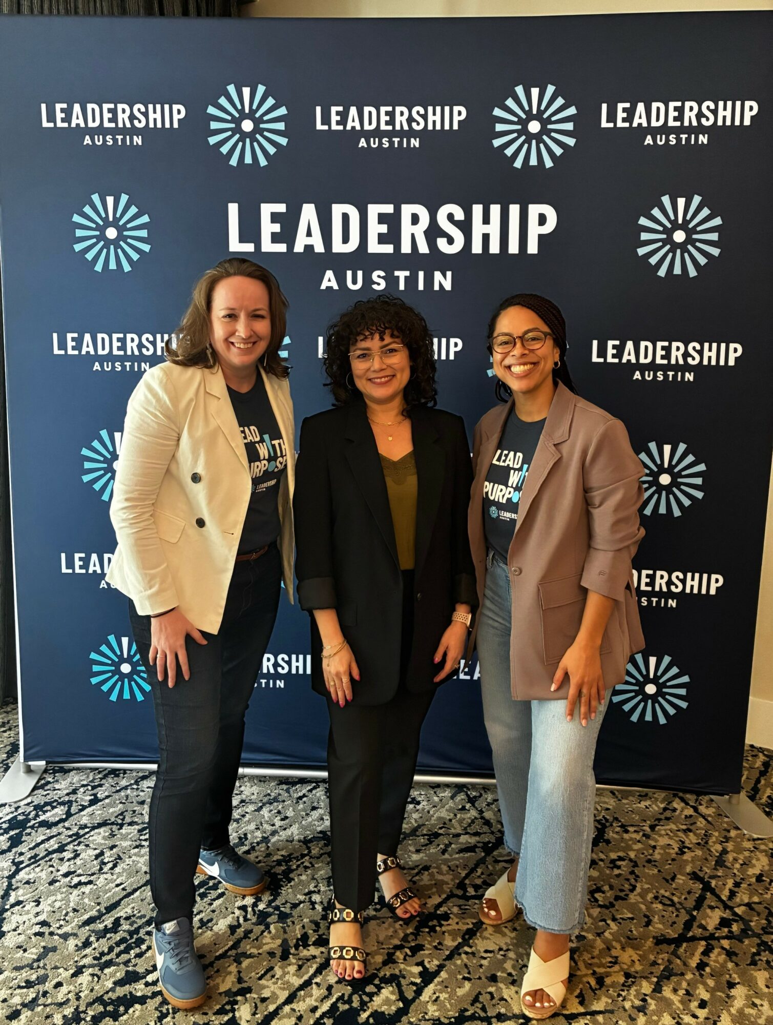 Three women standing in front of a blue backdrop with white text and logos, smiling, indoors. Word 'LEADERSHIP' is visible.