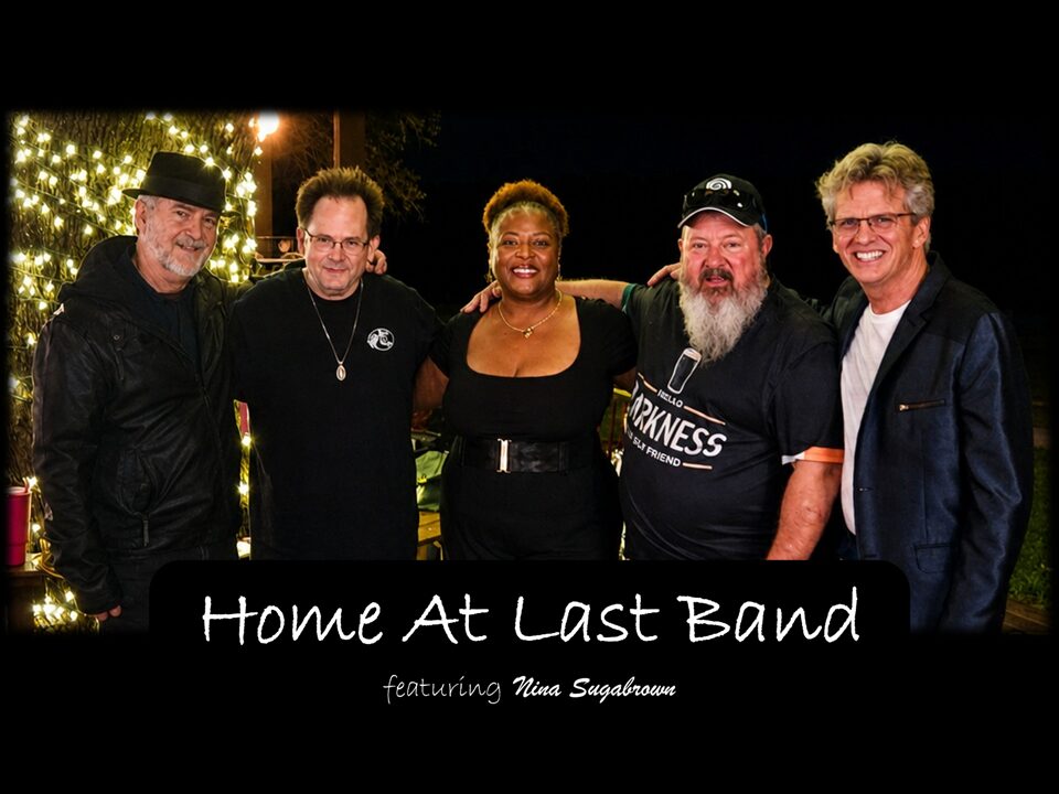 Five people standing together outdoors at night, smiling, with Christmas lights in the background. Text reads 'Home At Last Band' and 'featuring Nina Supernova'.