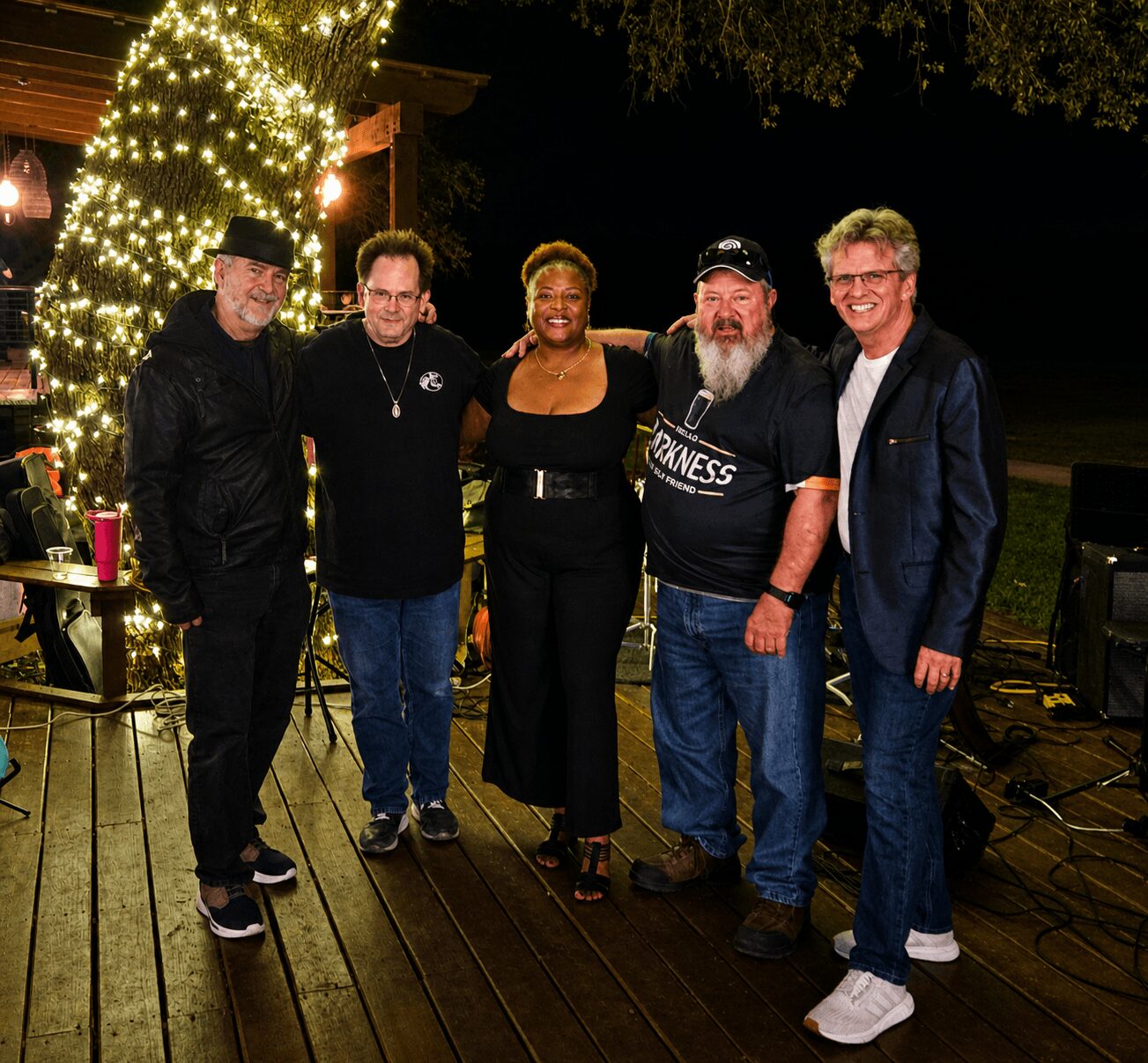 Group of five people standing outdoors at night, smiling, with a decorated tree and string lights in the background.