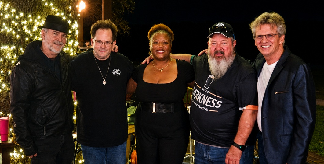 Group of five people standing outdoors at night, smiling, with Christmas lights in the background.
