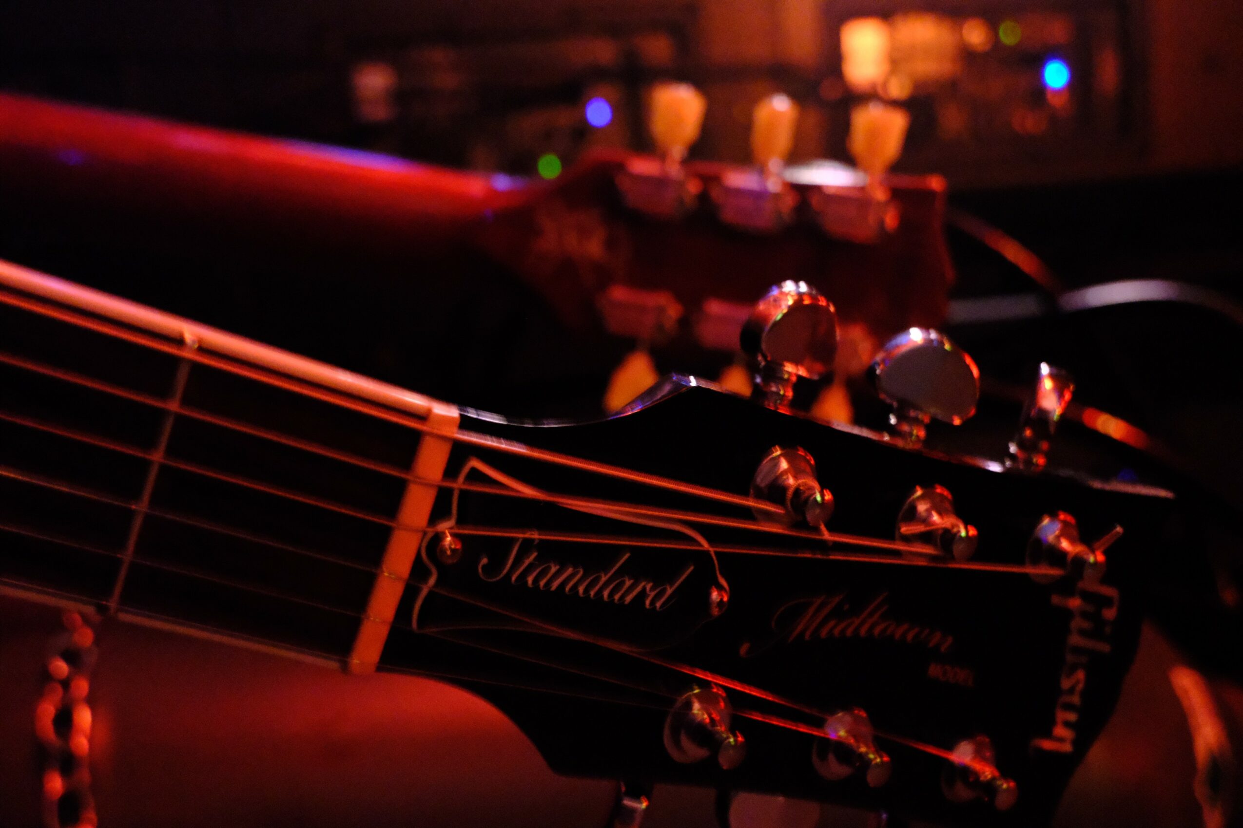 Close-up of a guitar headstock with tuning pegs and strings, with a blurred background of a music studio or stage, dimly lit.