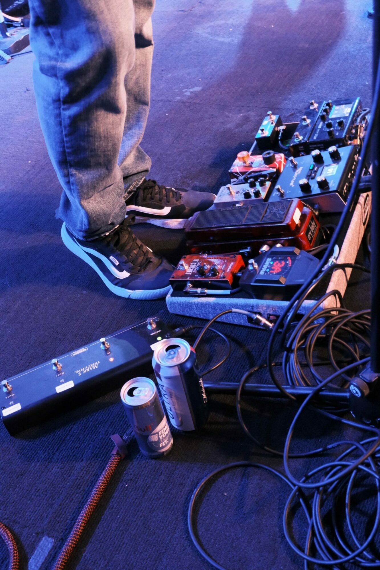 Person's legs and feet near a pedalboard with various guitar effects pedals, on a dark surface.