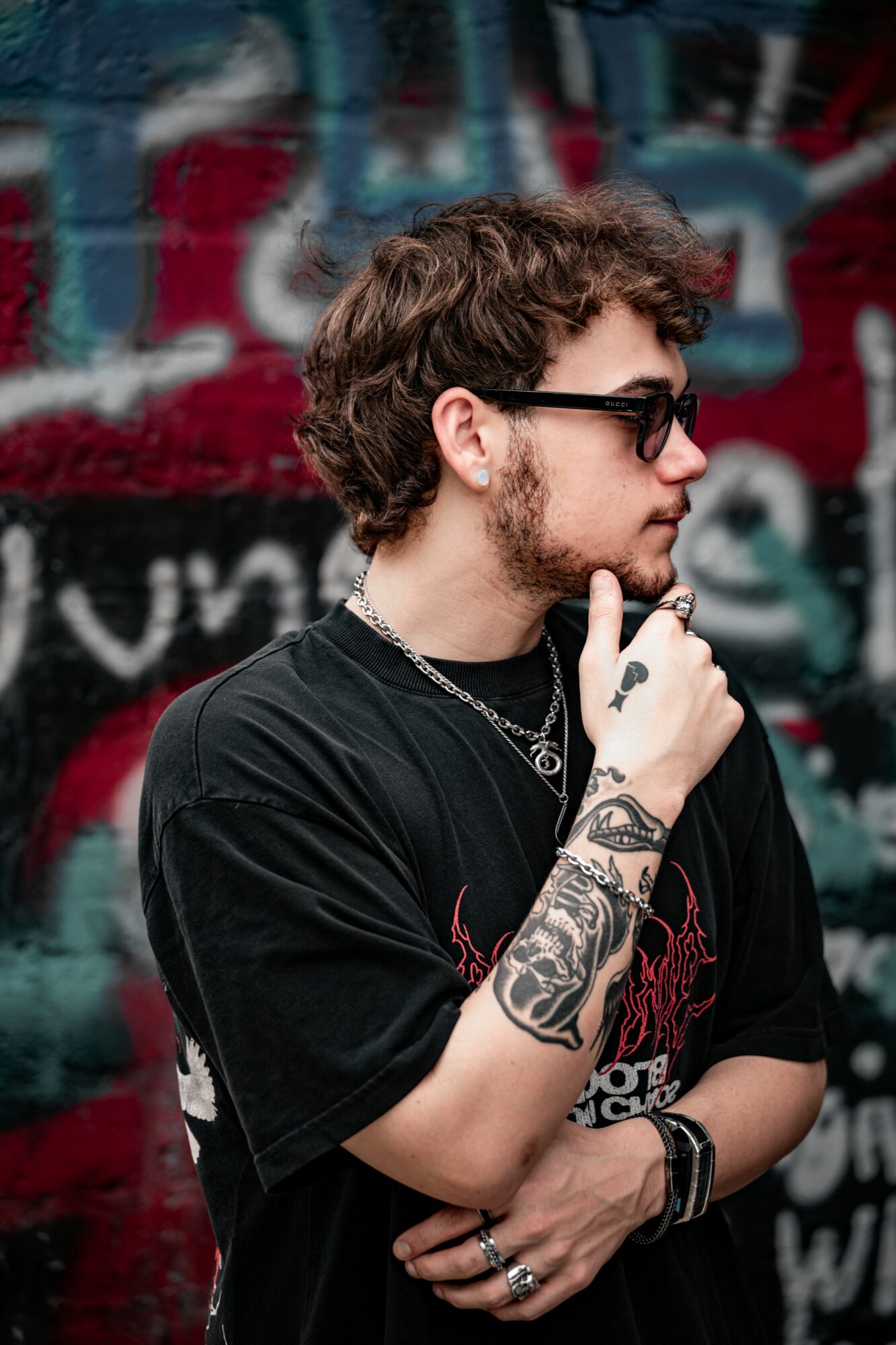 Young man with curly hair and glasses, wearing a black T-shirt, standing in front of a graffiti wall, with tattoos on his arm, holding his chin.