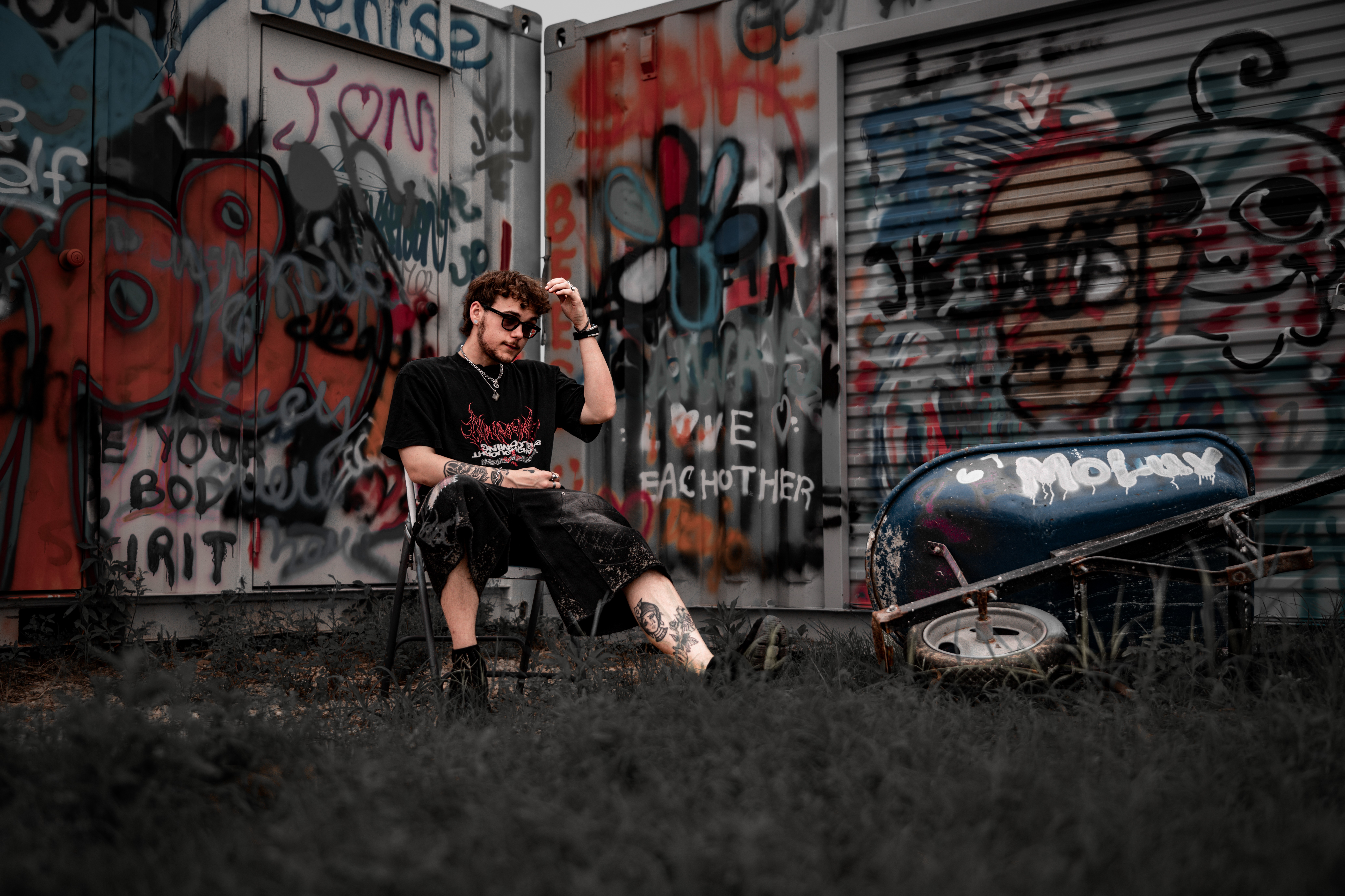 Person sitting on a chair in front of graffiti-covered wall, holding a drink, with a scooter nearby, outdoors.