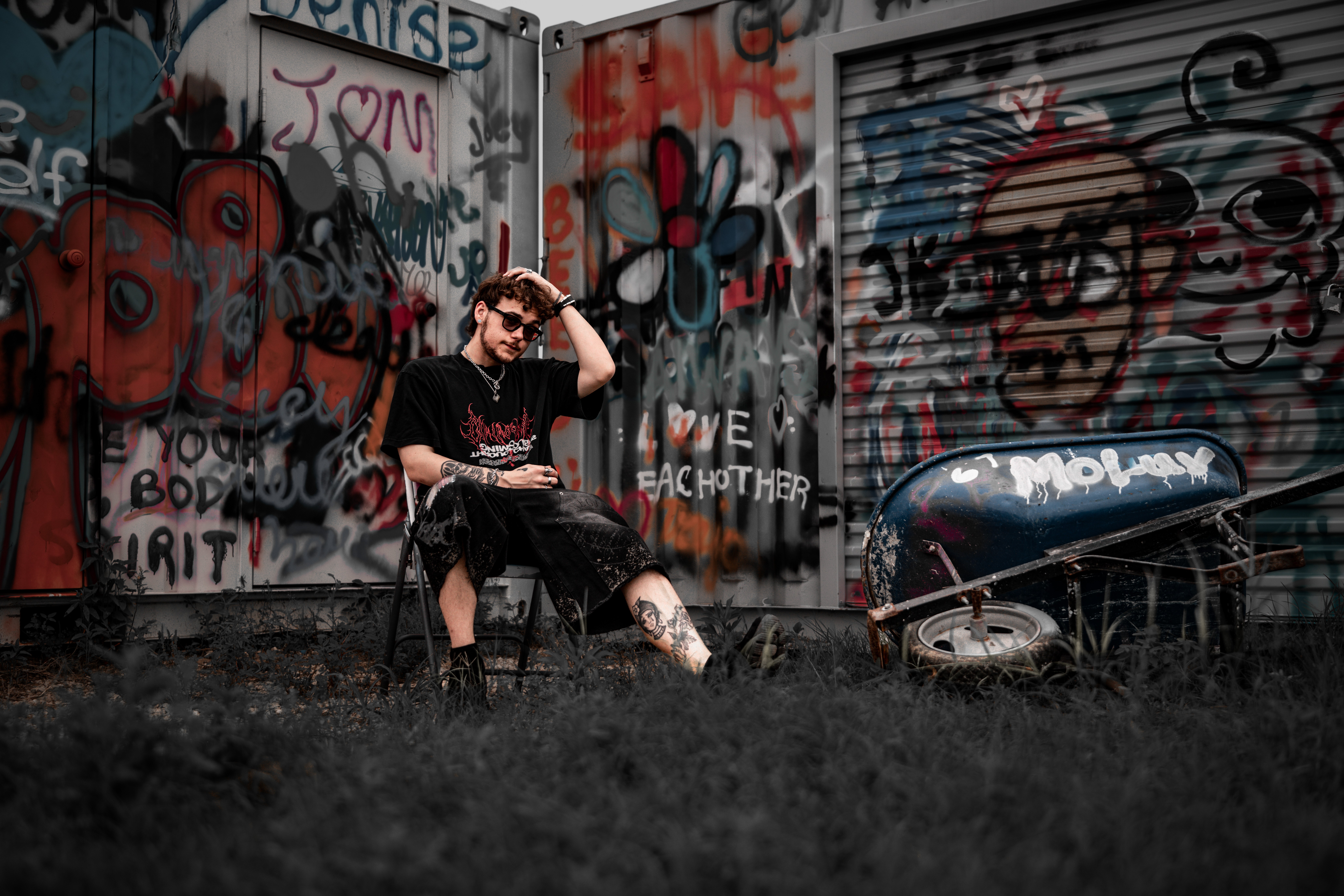 Person sitting on a chair in front of graffiti-covered wall, adjusting sunglasses, with a skateboard nearby.