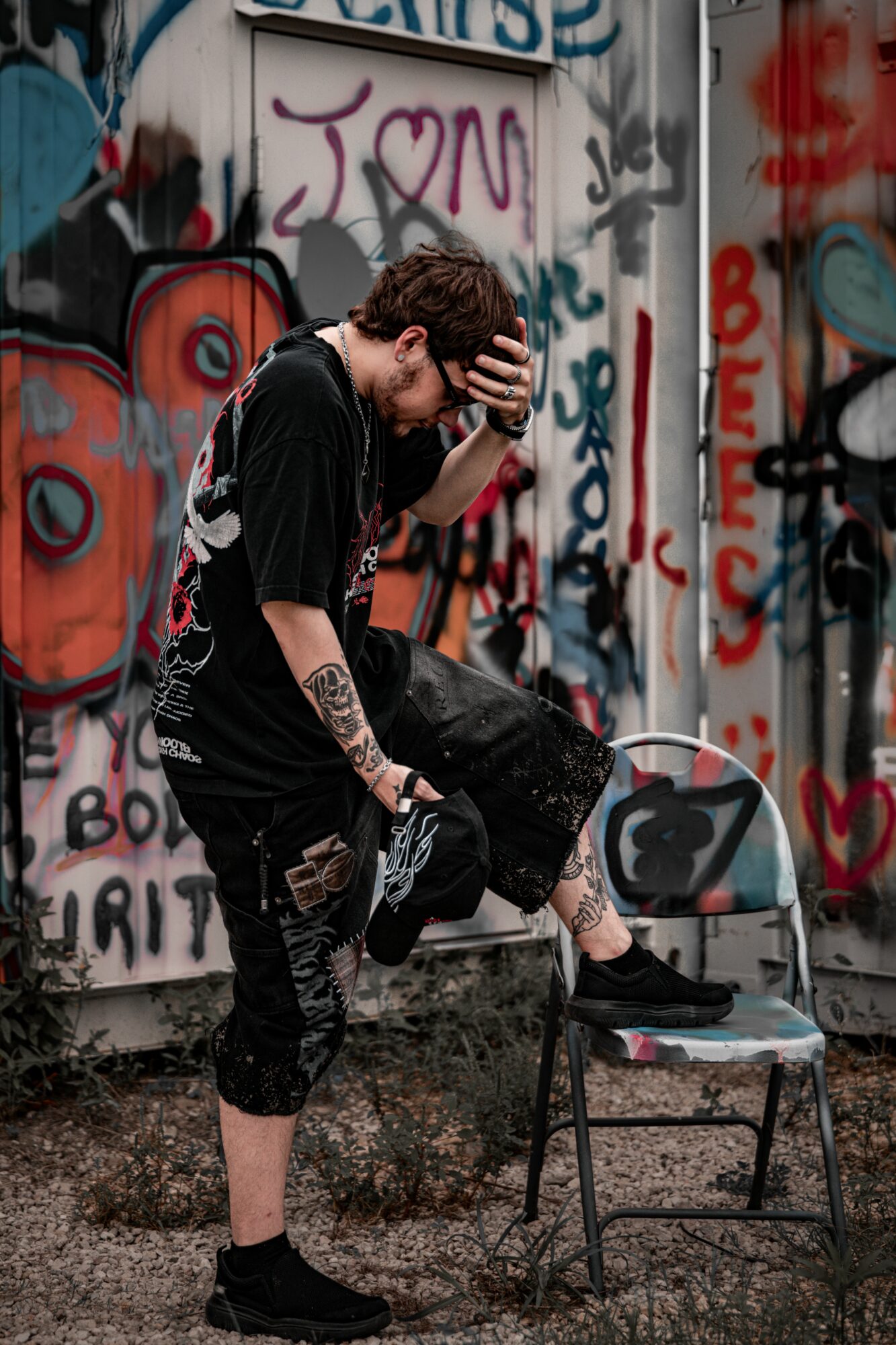 Young man with tattoos and dark clothing stands on one leg, holding his head, next to a chair in front of graffiti-covered wall.