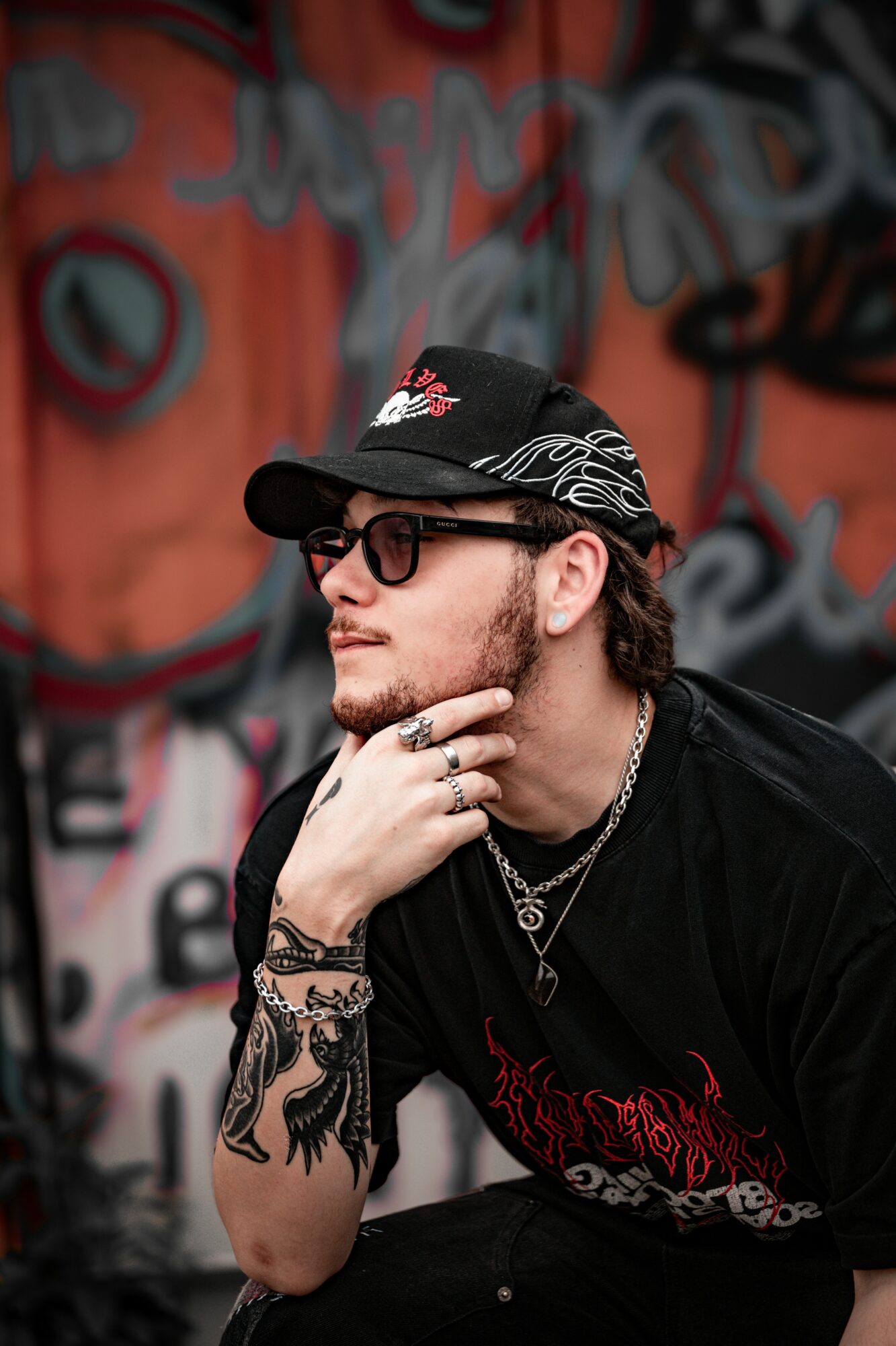 Young man with glasses, a cap, and tattoos, sitting thoughtfully against a graffiti wall.