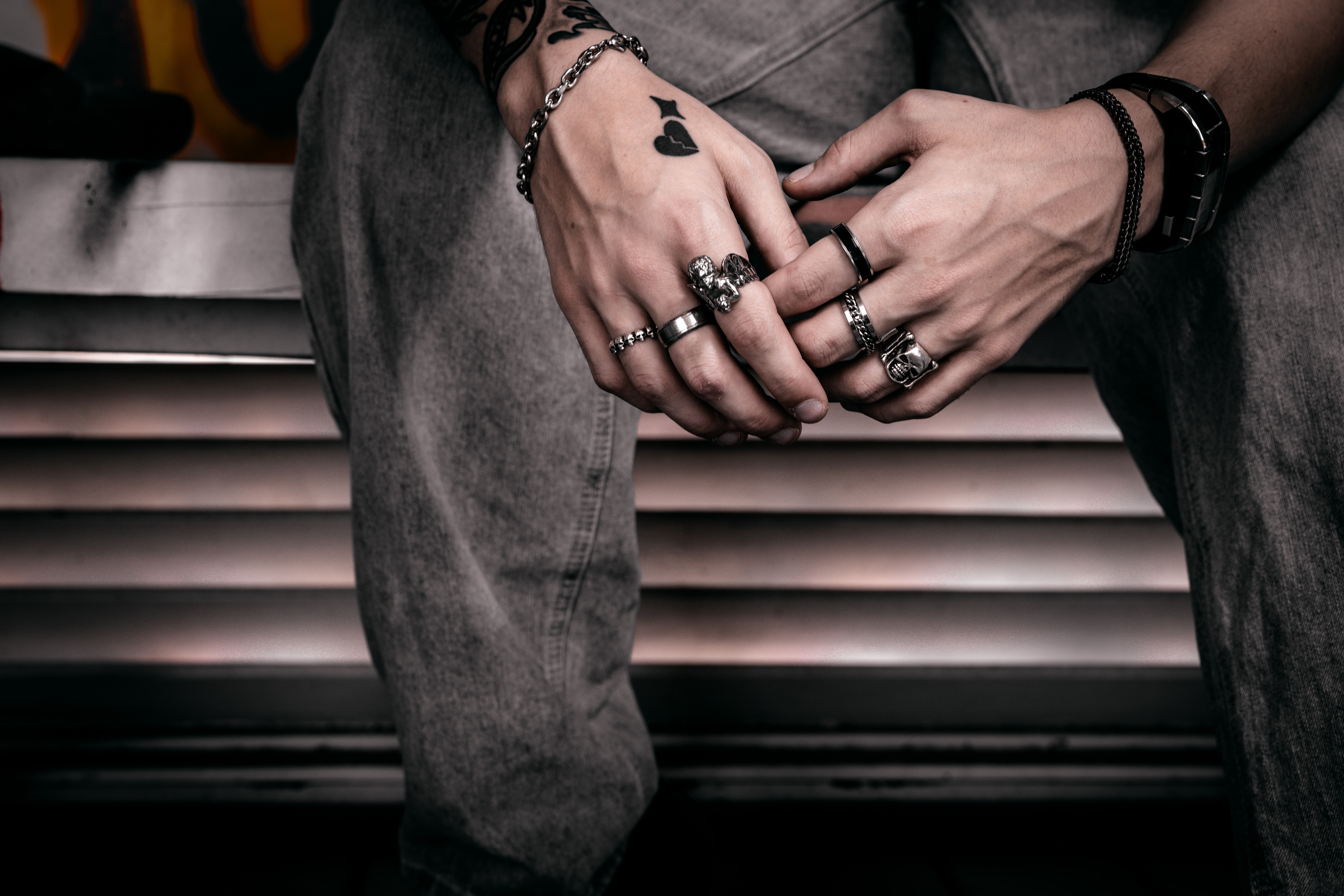 Person's hands with multiple rings and bracelets, resting on their lap, wearing dark clothing and sitting in front of a metallic background.