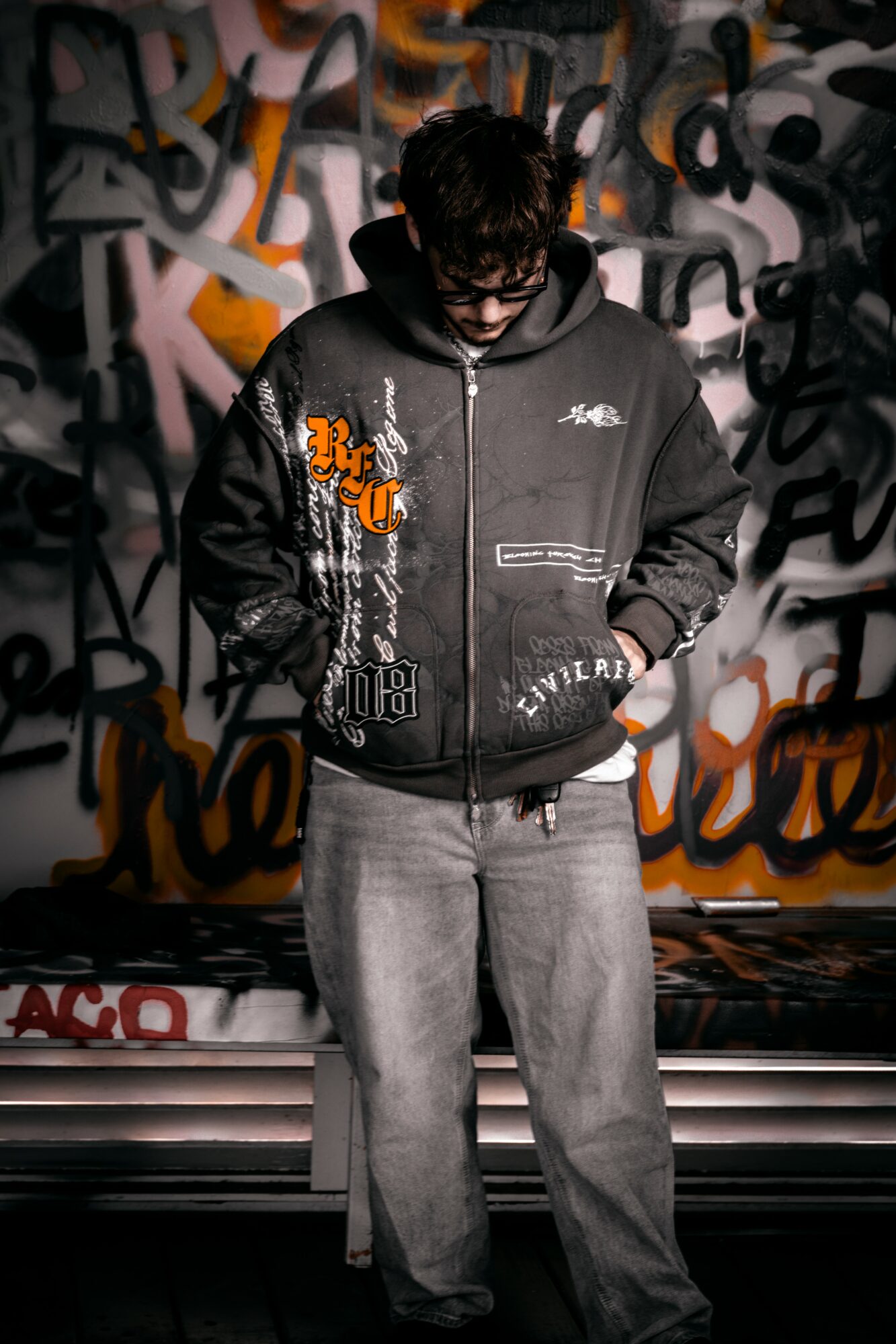 Person with dark hair and glasses stands in front of graffiti wall, wearing a black jacket and gray pants.