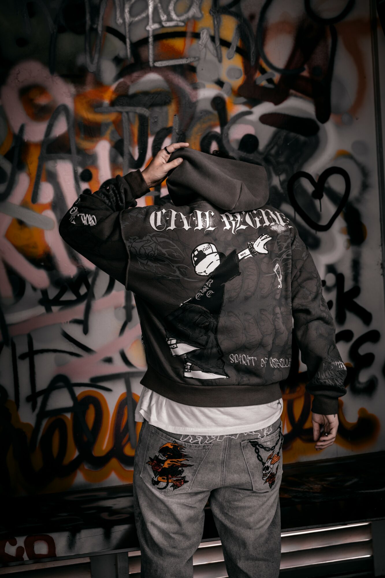 Person wearing a black hoodie with graffiti, standing in front of a graffiti wall, face hidden, hand on head.