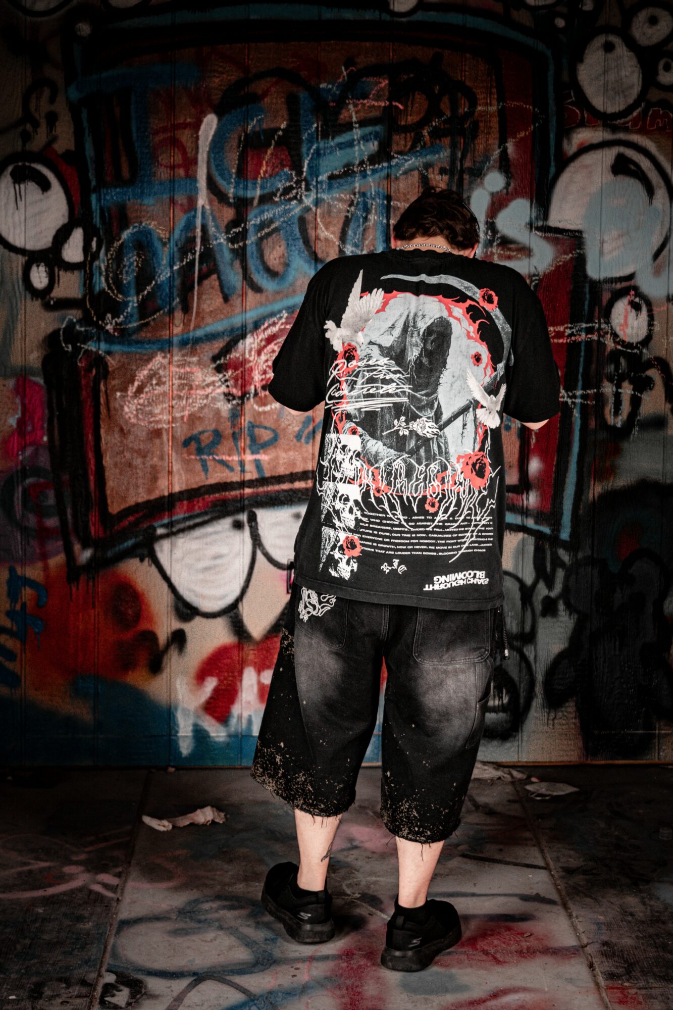 Person standing with back to camera in front of graffiti wall, wearing a black and white graphic shirt and loose pants.