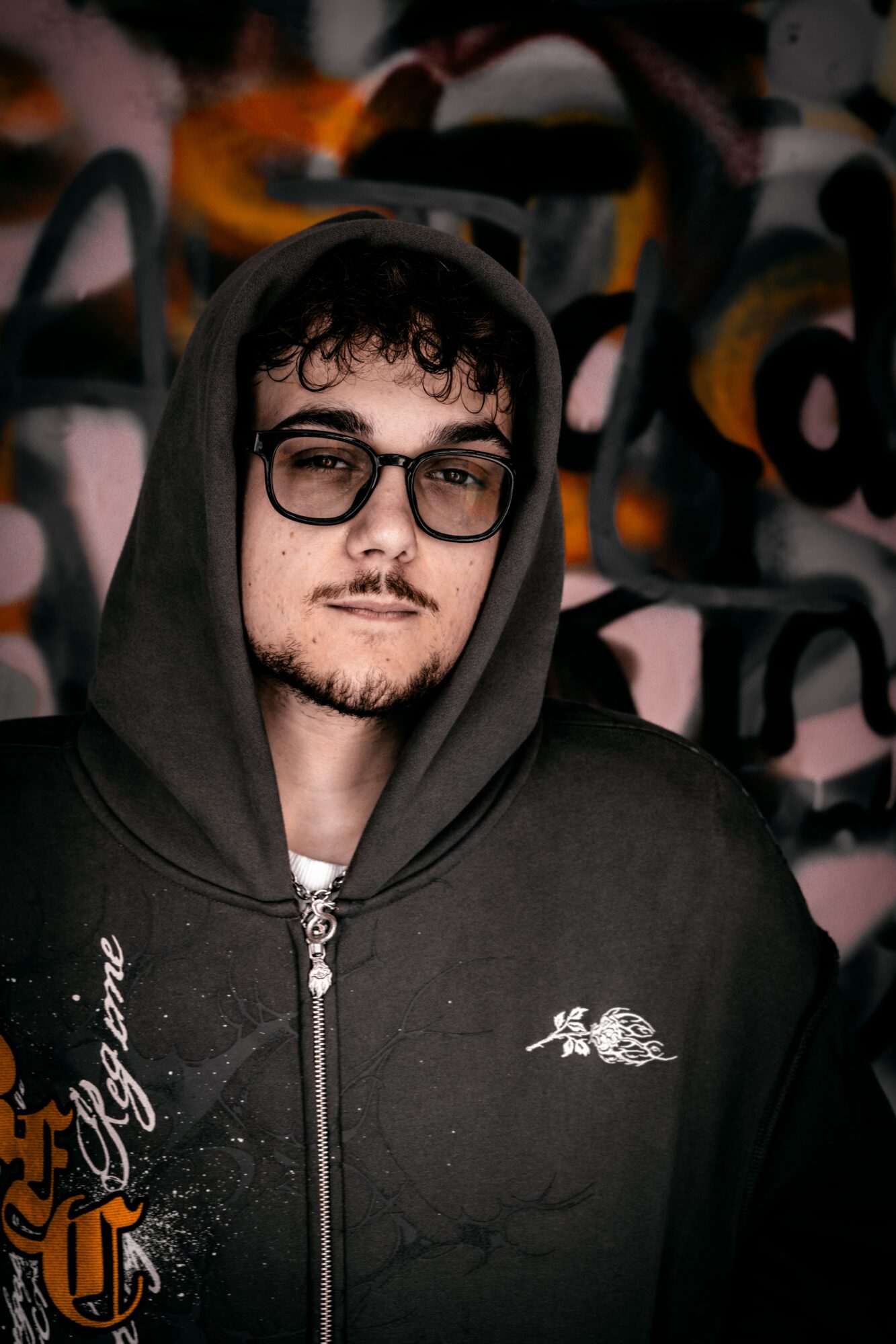 Young man with glasses and curly hair wearing a black hoodie, standing in front of a colorful graffiti wall.