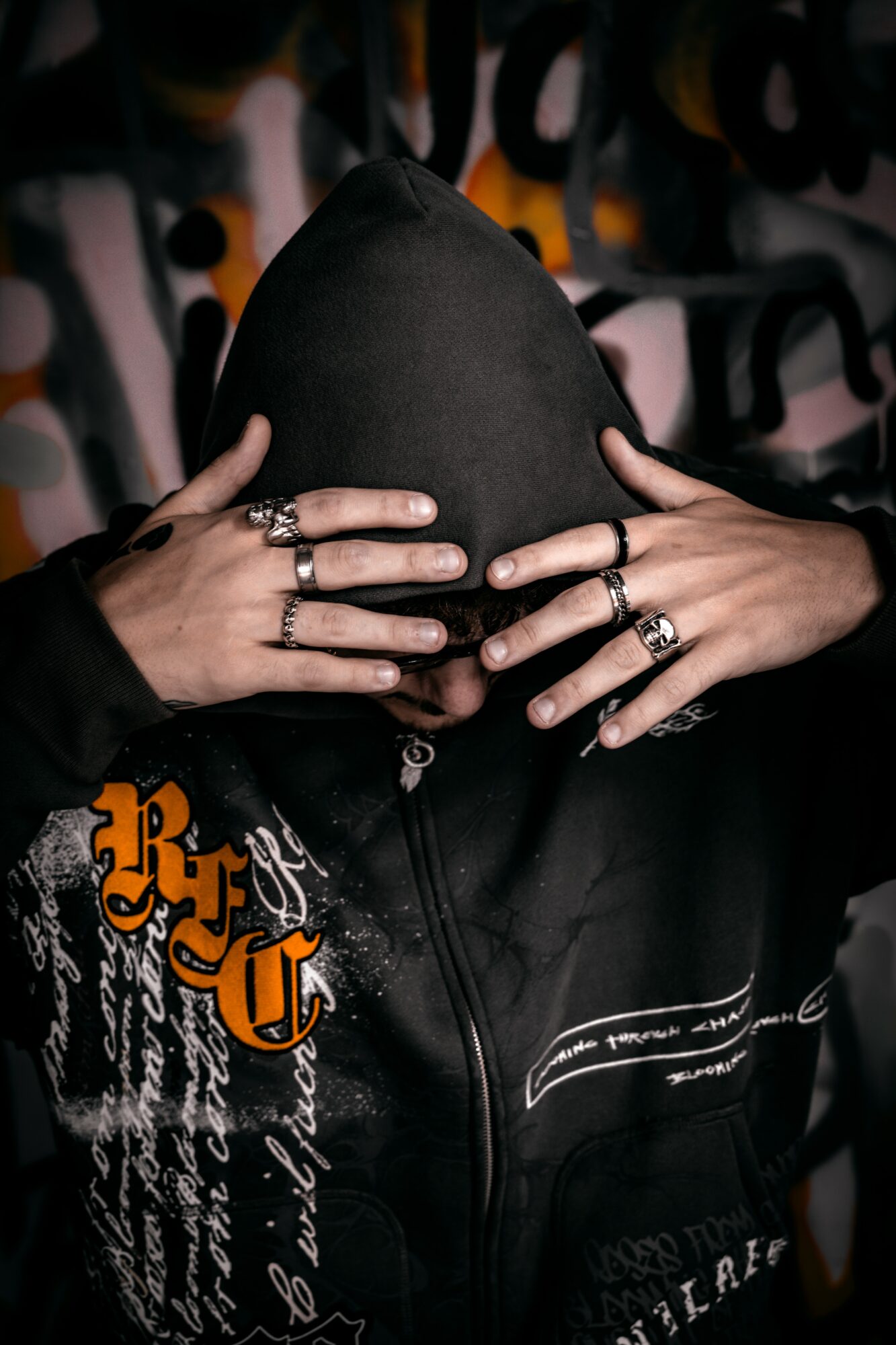 Person wearing a black hoodie covering face, showing multiple rings on fingers, with graffiti background.