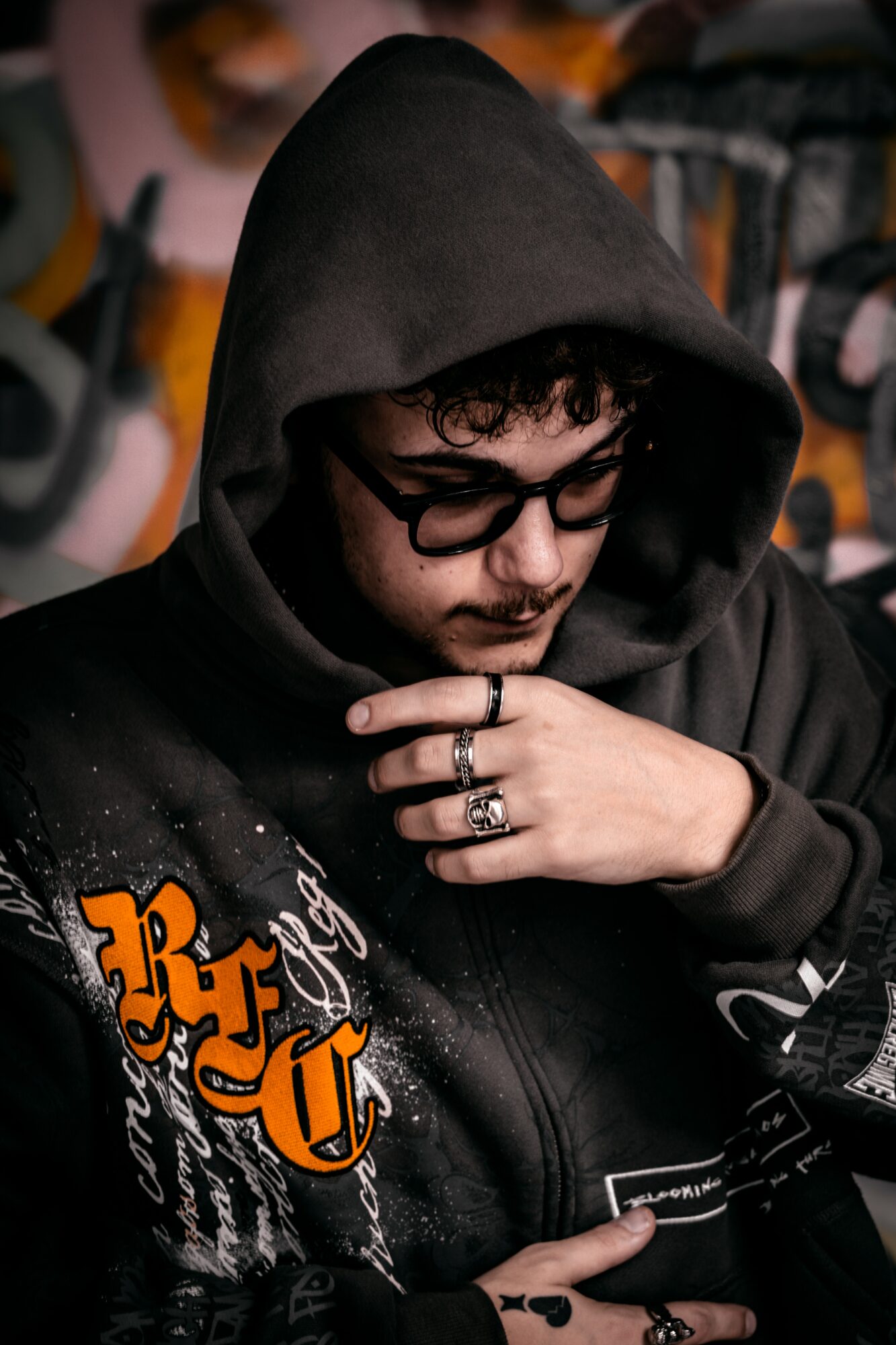 Young man with glasses wearing a black hoodie, looking down with hand near face, graffiti background.