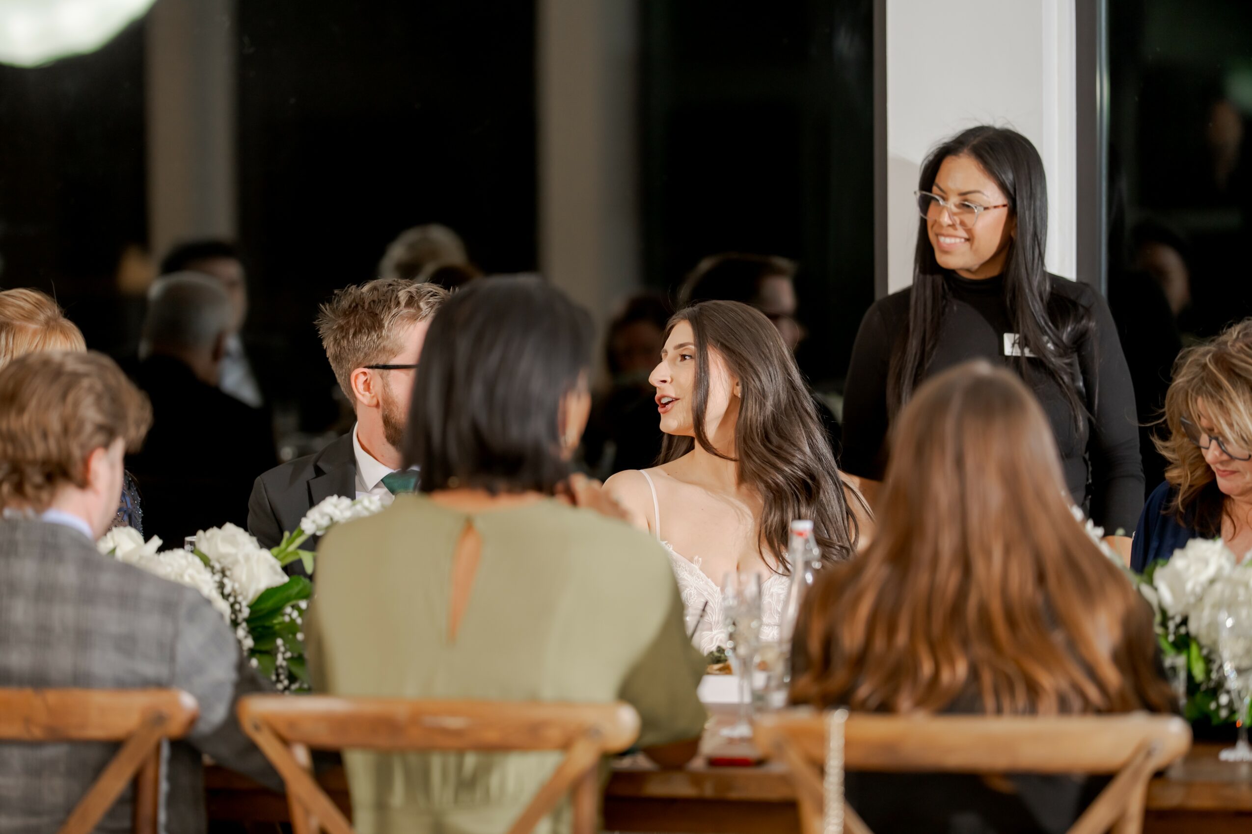 Group of people seated at a table during a social event, with a woman standing and smiling nearby.