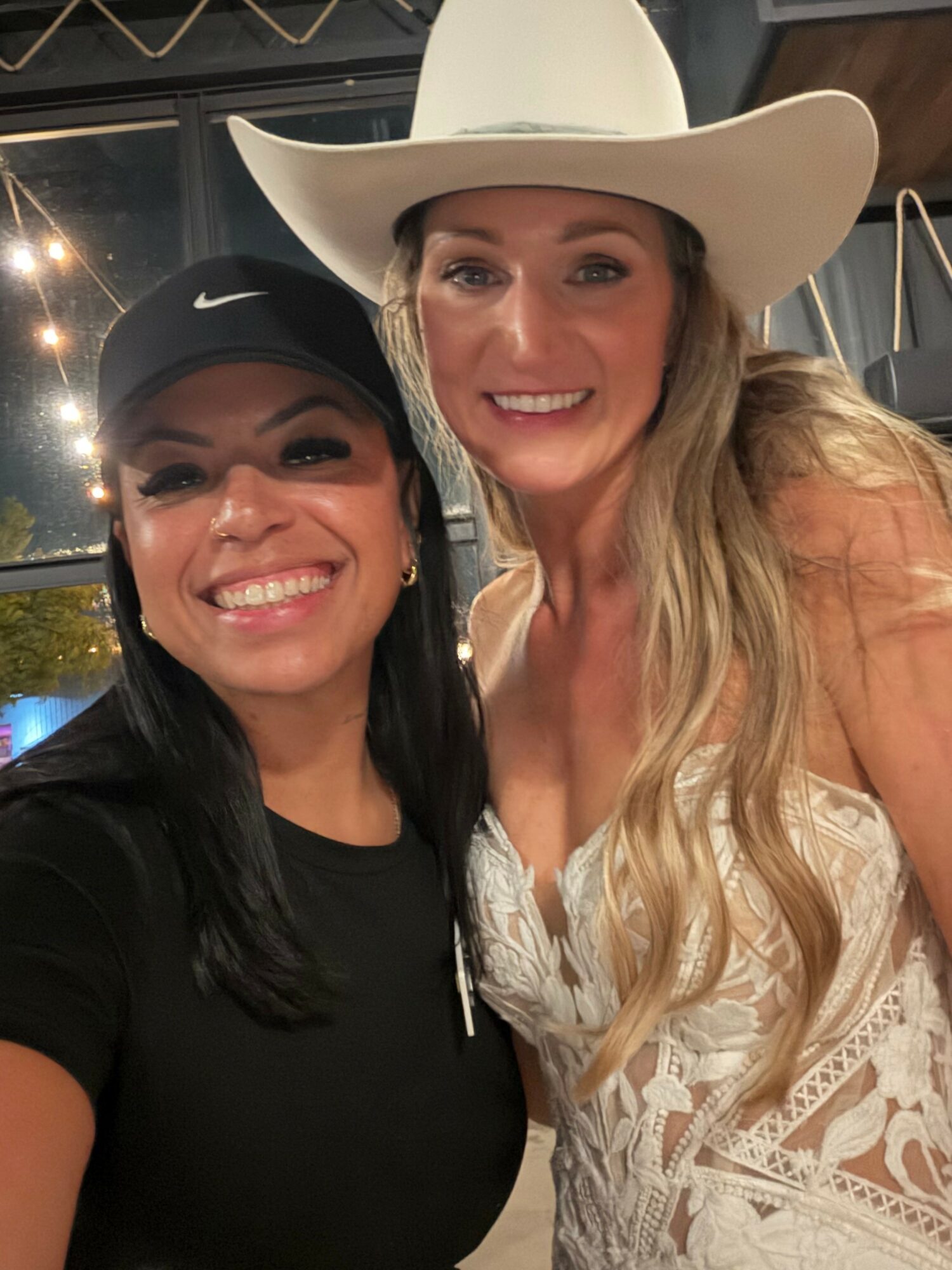Two women smiling, one wearing a cowboy hat and the other a black cap, indoors at night.