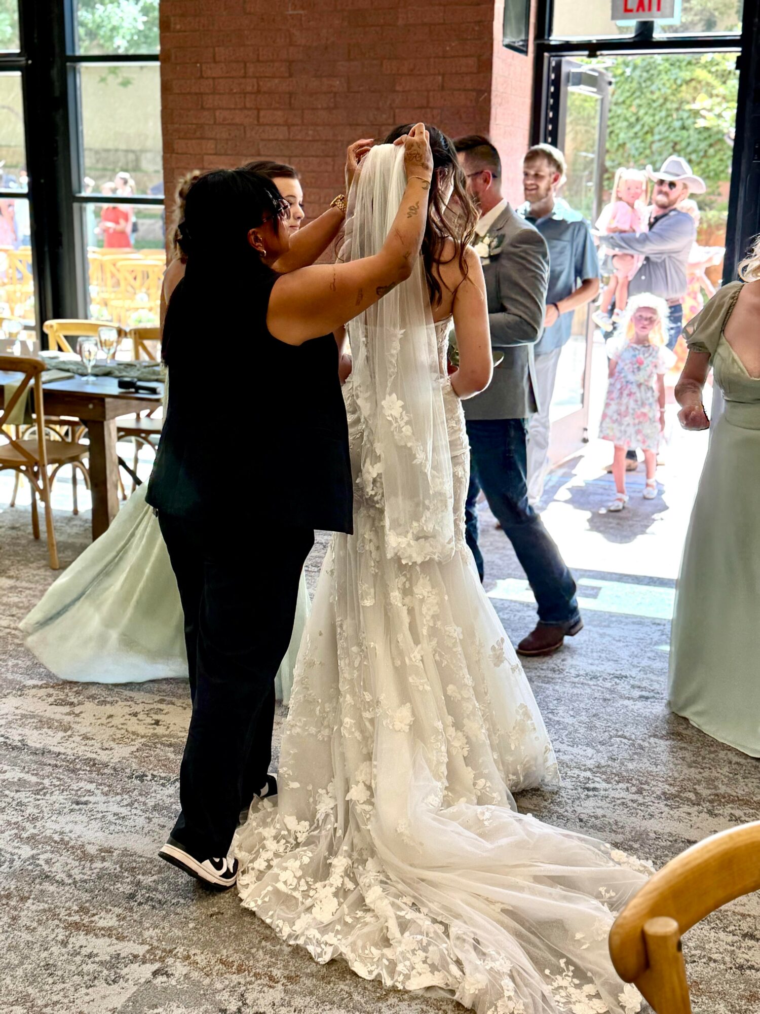 Person placing a crown on a bride's head during a wedding ceremony inside a venue with large windows and guests outside.