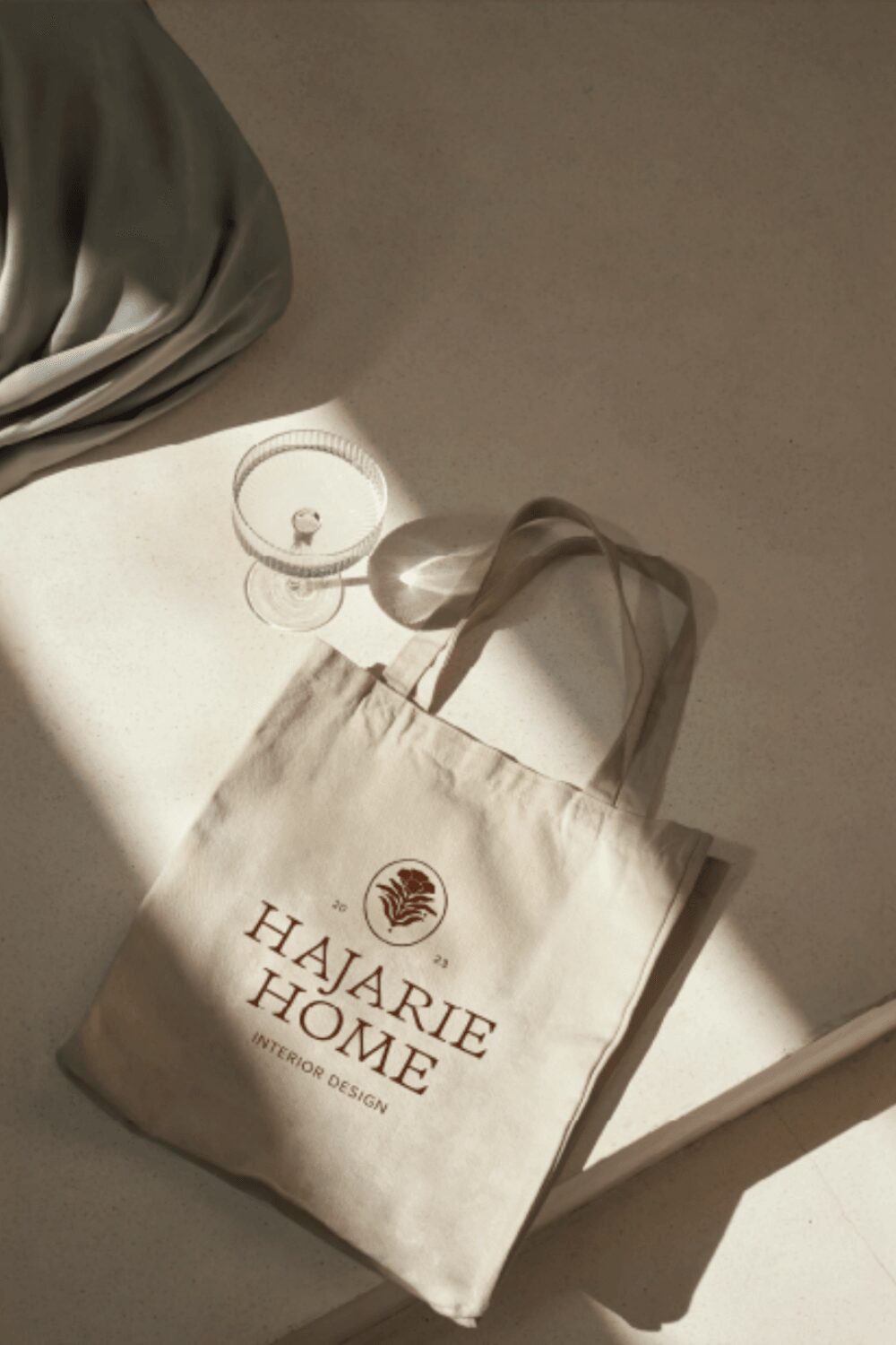 Canvas bag with 'Haarie Home' logo and text, lying on a surface with a shadow, next to a glass and fabric, with a tag attached.