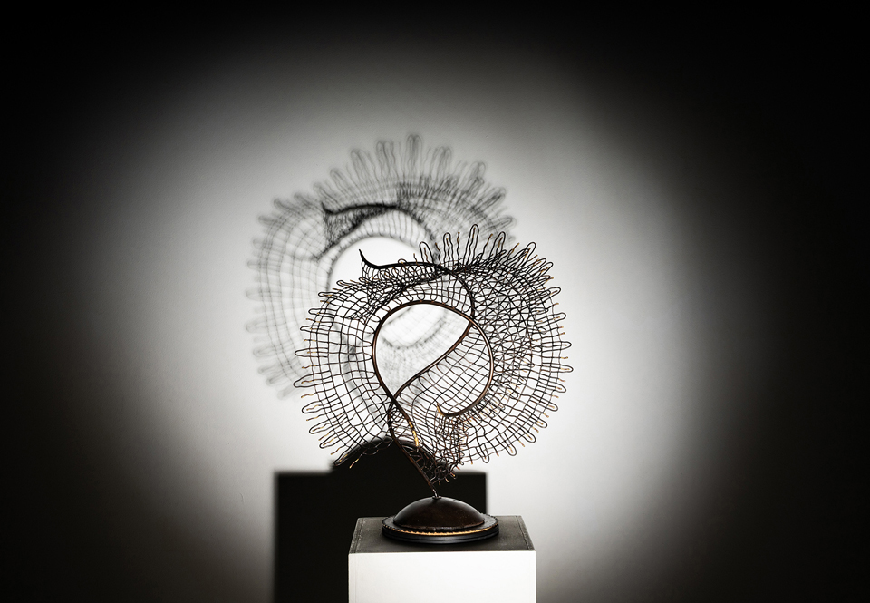 Abstract wire sculpture with circular and spiral shapes on a pedestal, illuminated against a dark background.