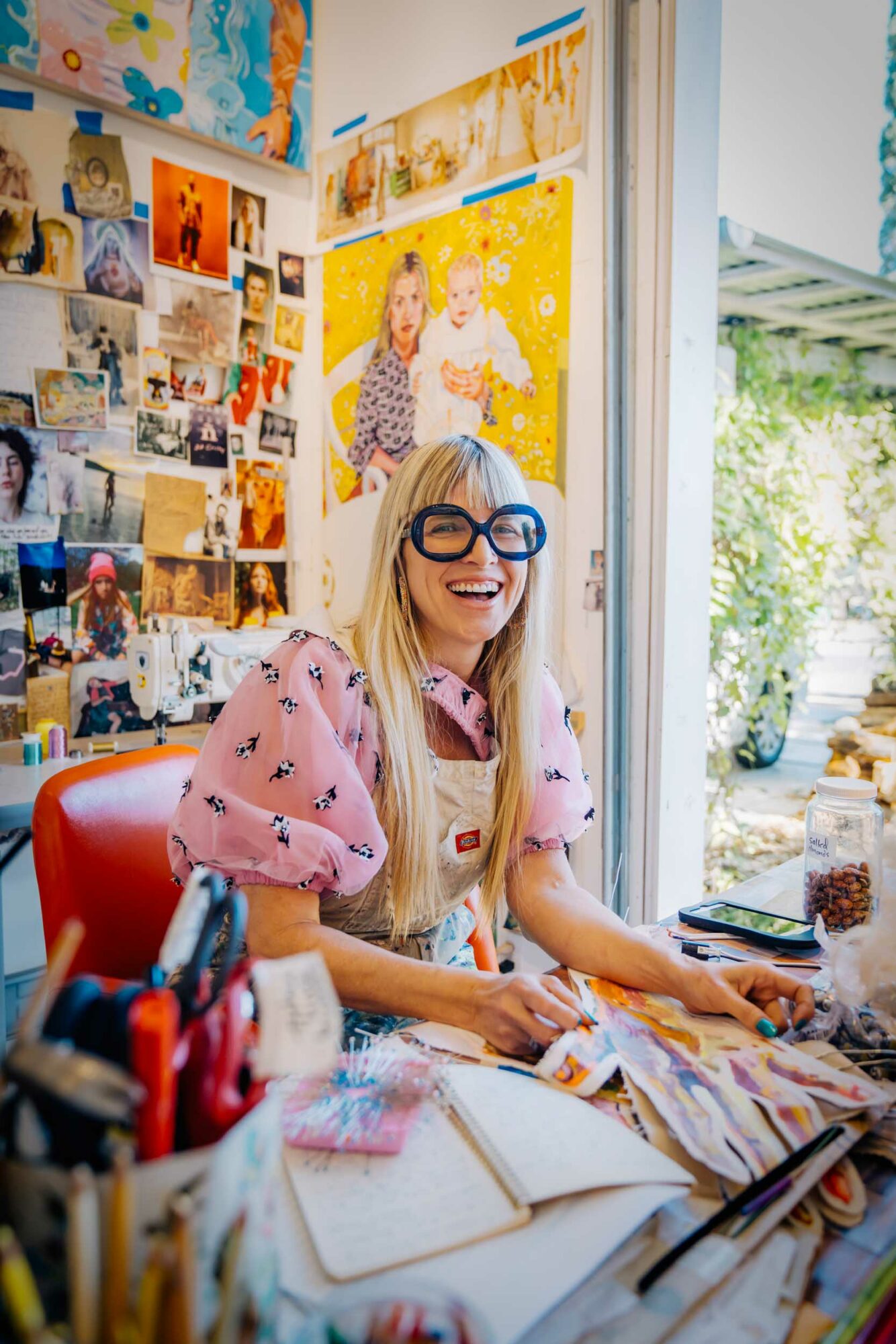 Woman with glasses smiling at a desk surrounded by fabric and art supplies, with colorful artwork on the wall behind her.