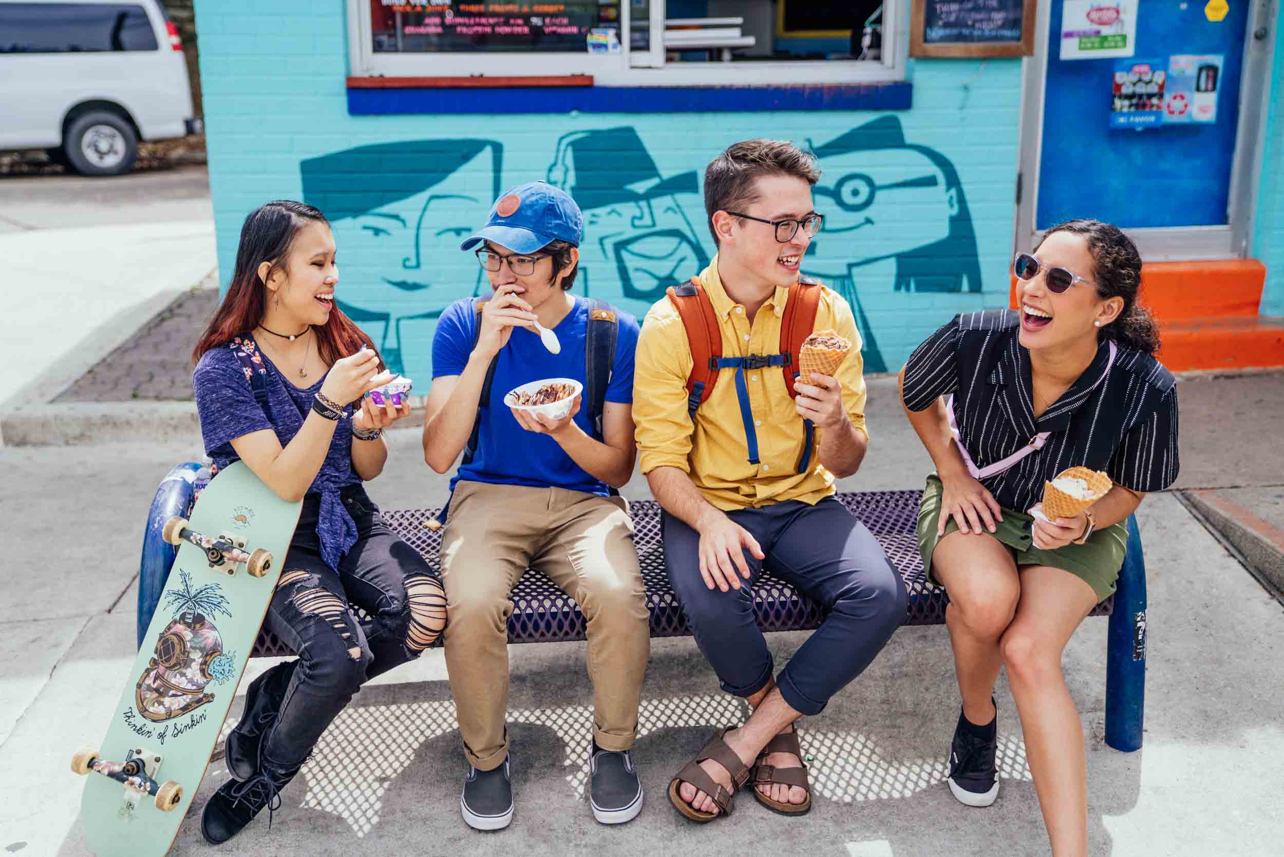 Four young people sit on a bench eating ice cream outside a colorful building, smiling and chatting.