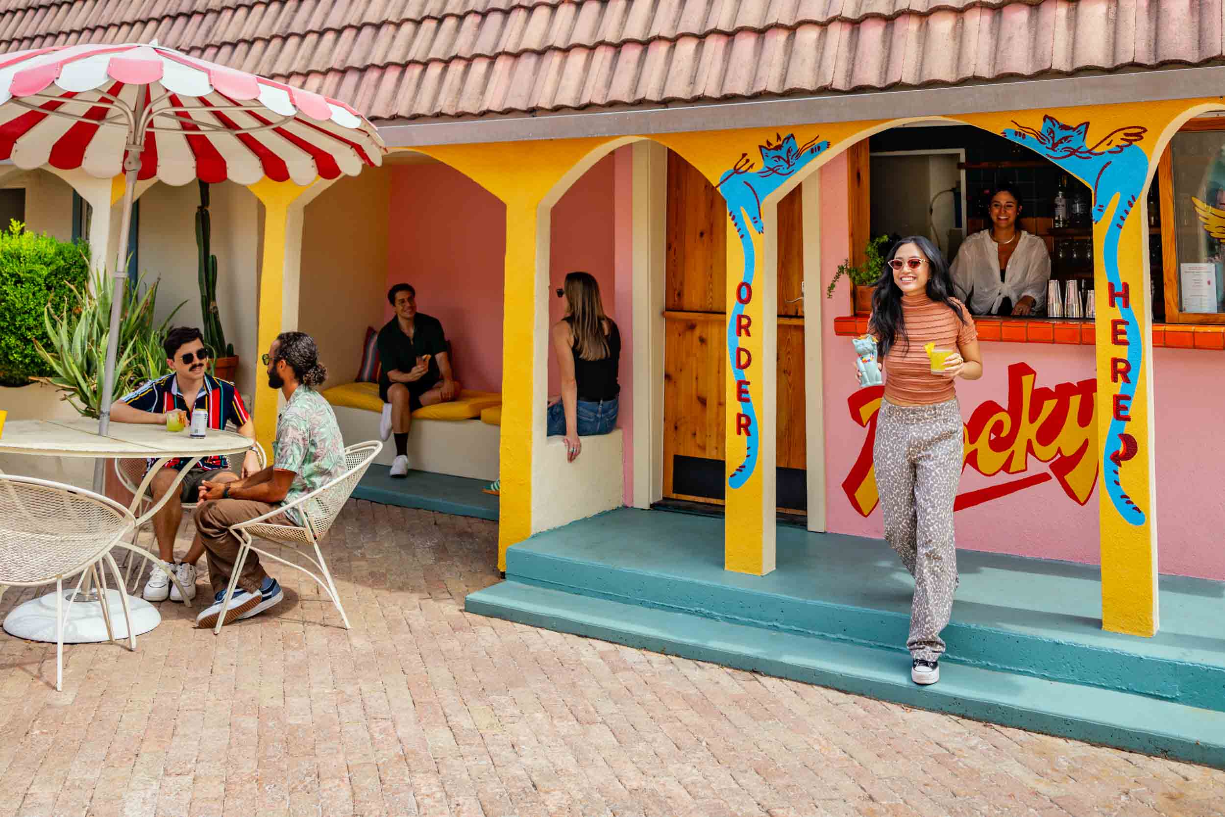 People sitting outside a colorful ice cream shop with a woman standing in front, under a striped umbrella, on a brick patio.