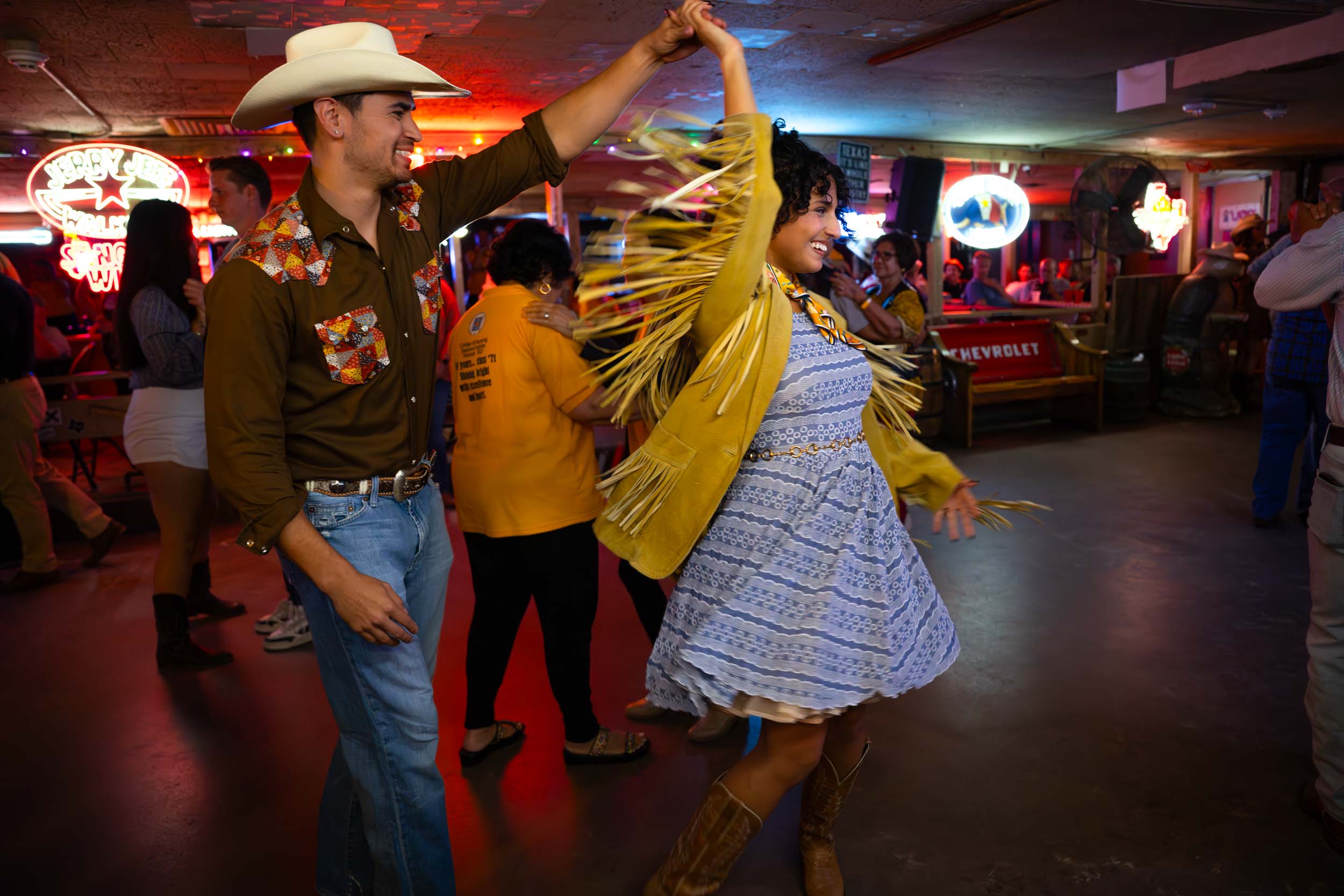 Two people dancing in a bar, one wearing a cowboy hat and the other in a yellow jacket, with a lively background.