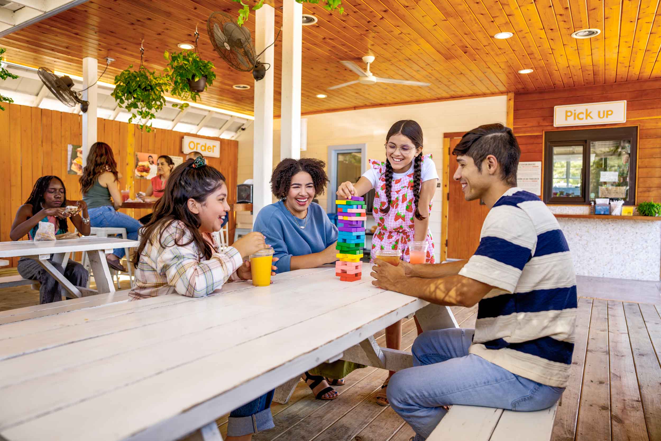 Four people playing a colorful stacking game at a table in a bright, wooden interior space with other people in the background.