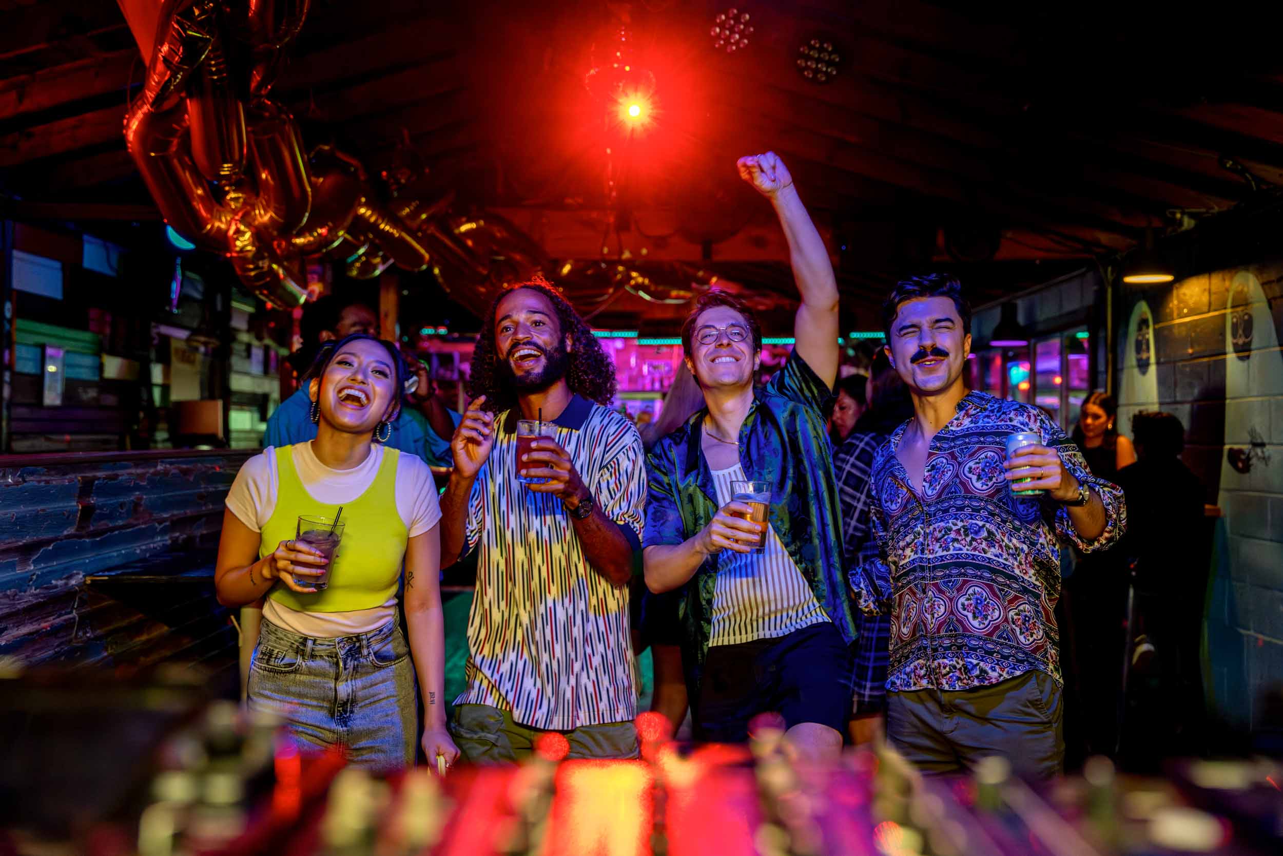 Four people celebrating with drinks in a lively bar, some raising their fists, colorful lights in background.