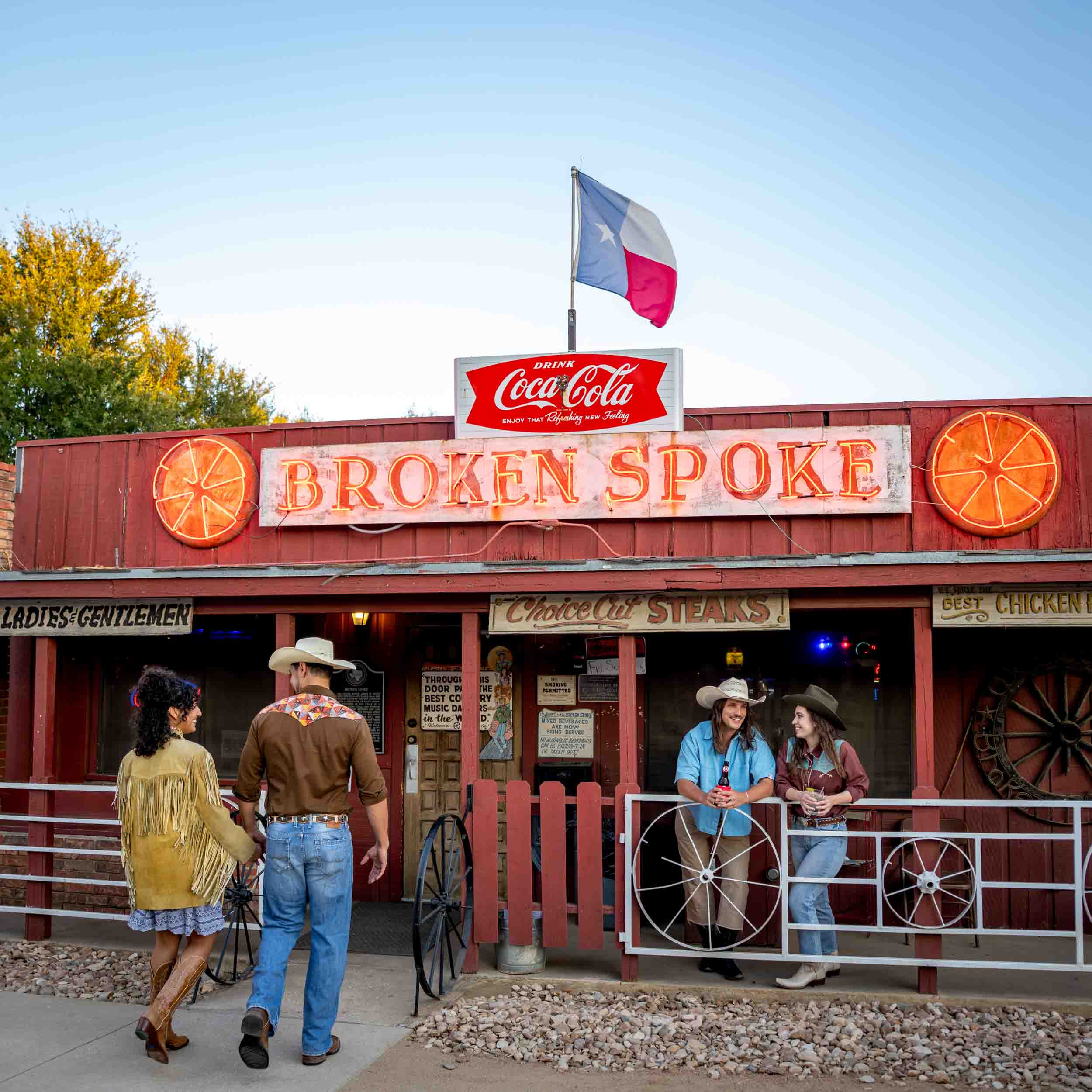 People walk past a Western-style building with a sign reading 'Broken Spoke' and a Texas flag on top.