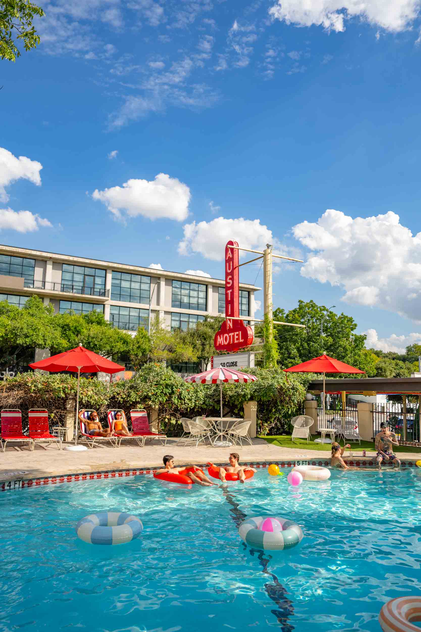 People swimming in a pool with red umbrellas and a hotel sign in the background, under a blue sky with clouds.