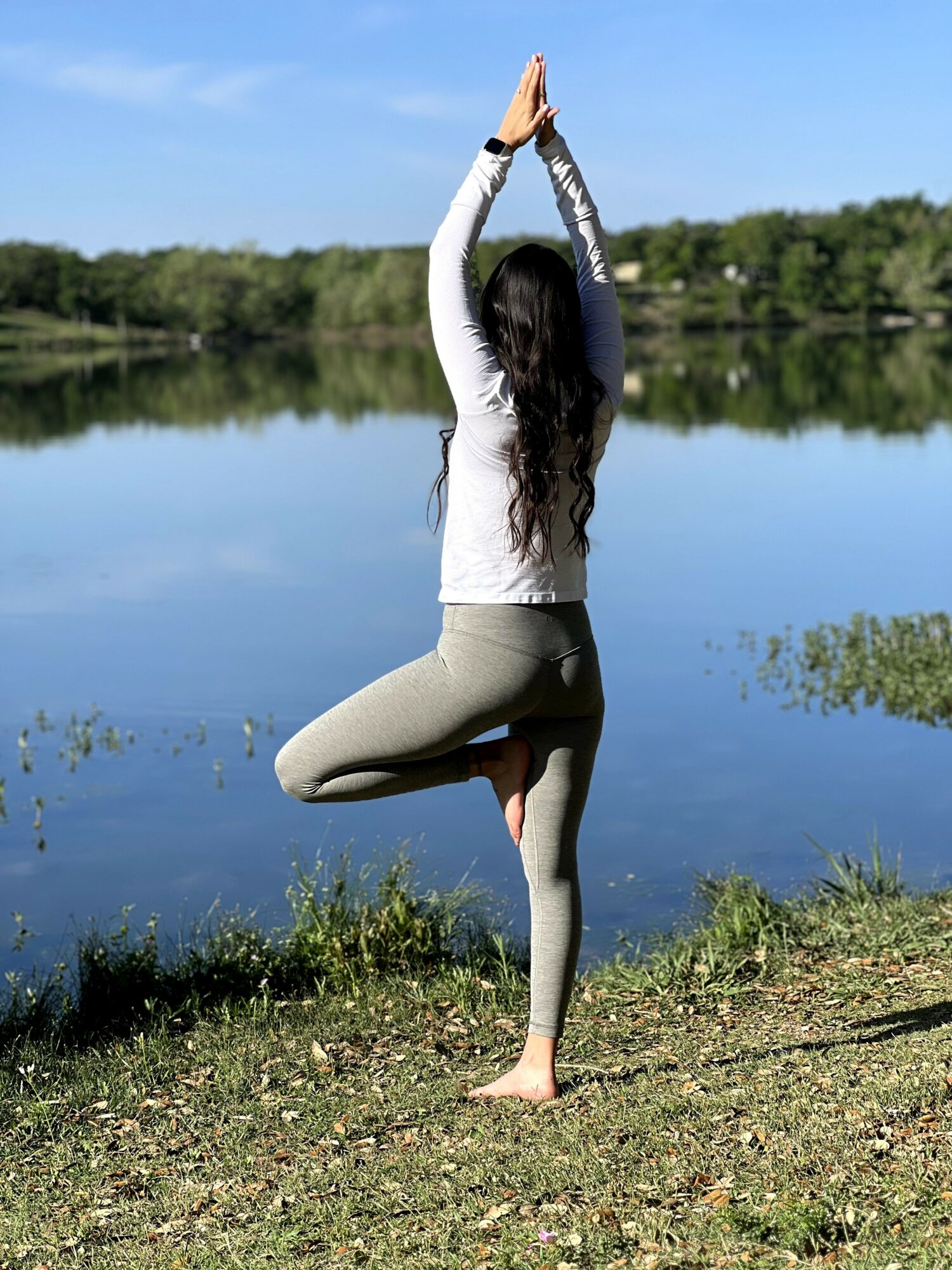 Person practicing yoga outdoors near a lake, standing on one leg with hands raised above head in a tree pose.
