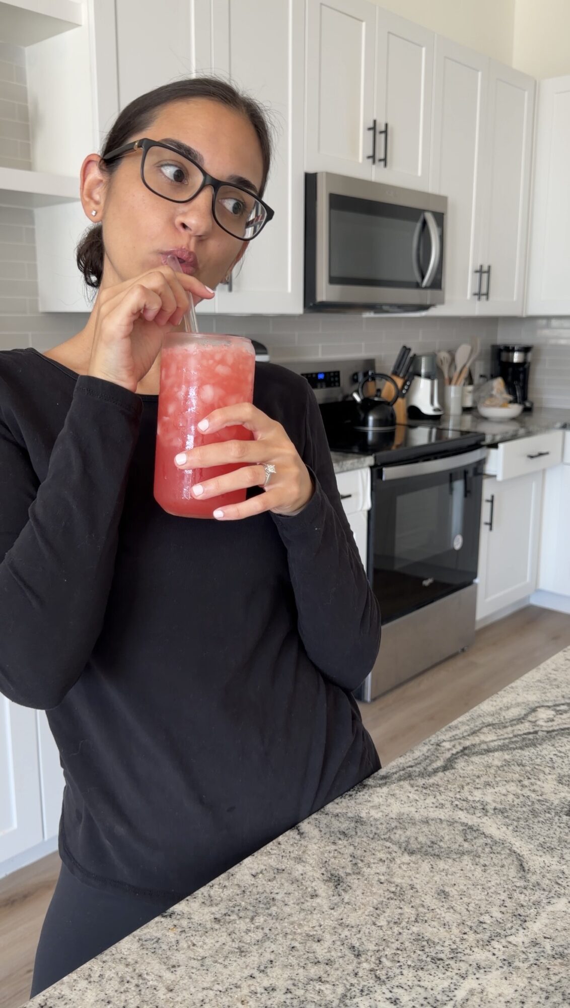 Woman with glasses drinking a red beverage from a glass in a kitchen.