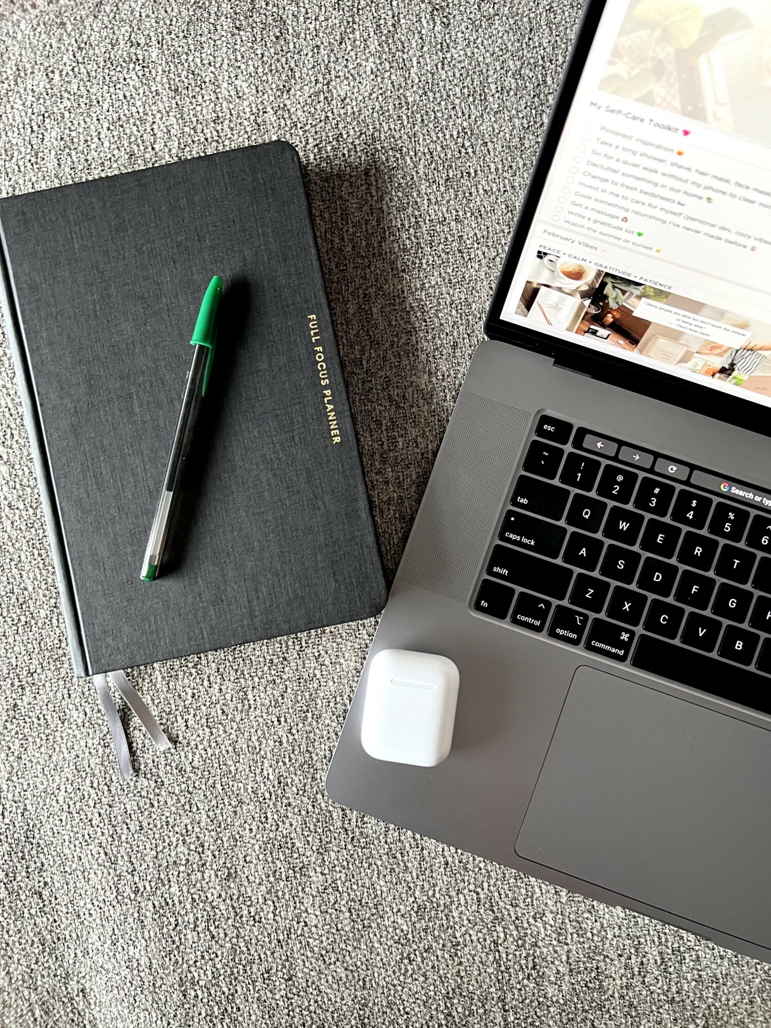 Laptop with open screen, gray notebook with pen on top, and white AirPods case on gray carpet.