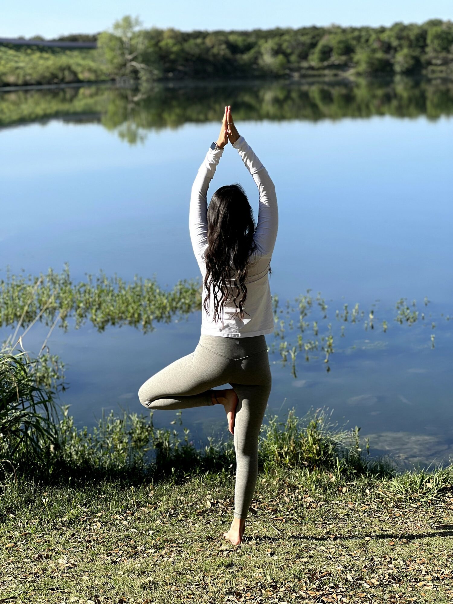 Person practicing yoga in tree pose near a lake with trees and sky reflected in water.