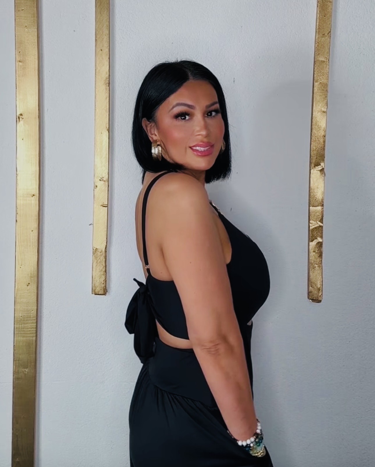 Woman with black hair and makeup, wearing a black dress with thin straps, standing against a white wall with gold accents.