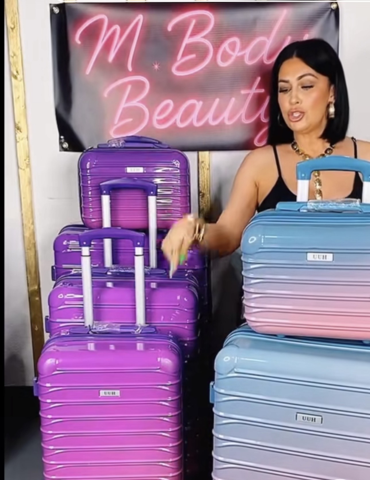 Woman with black hair and earrings stands next to stacked purple and blue suitcases, in front of a pink neon sign that reads 'M Body Beauty'.