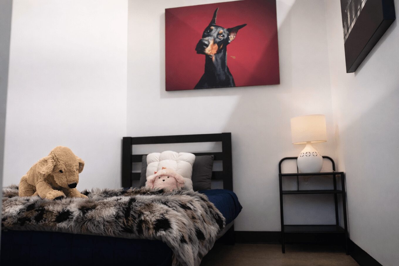 A bedroom with a bed, stuffed animals, a painting of a dog, and a nightstand with a lamp.
