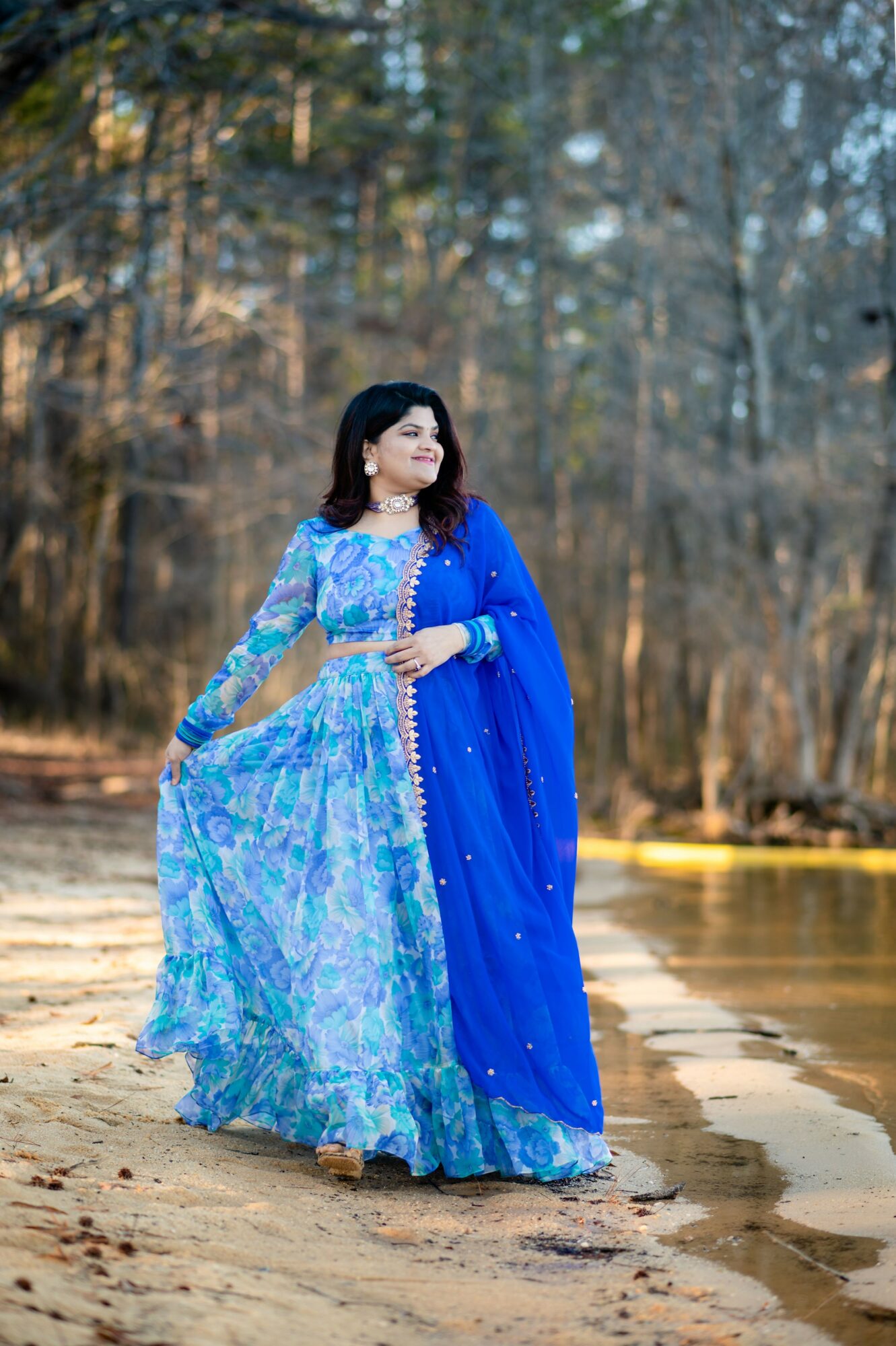 Woman in a blue and turquoise dress walking on a sandy path near water with trees in the background.