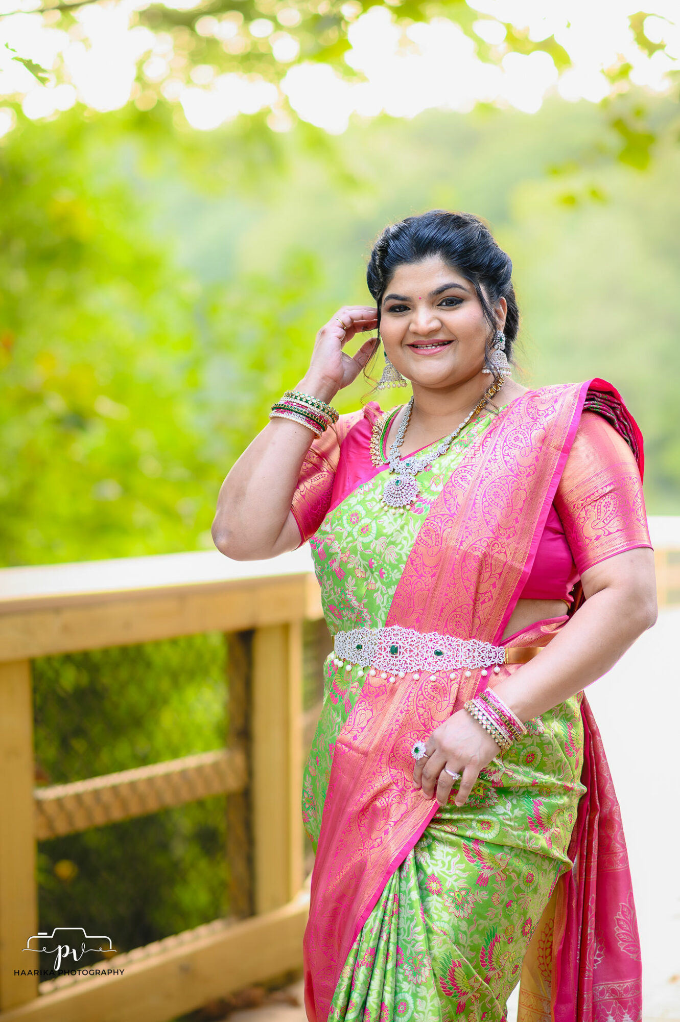 Smiling woman in colorful traditional attire standing outdoors on a wooden bridge with green foliage background.