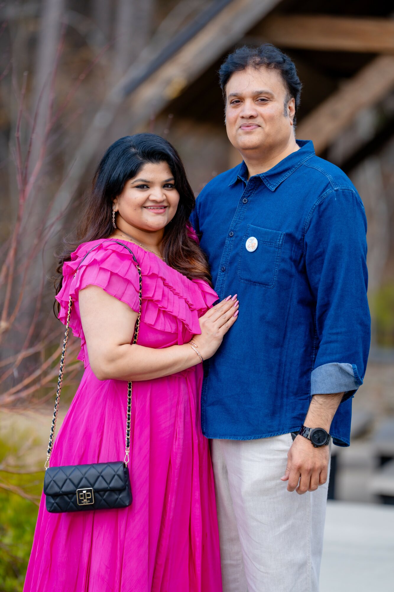 A smiling woman in a pink dress and a man in a blue shirt stand outdoors, with trees and a wooden structure in the background.