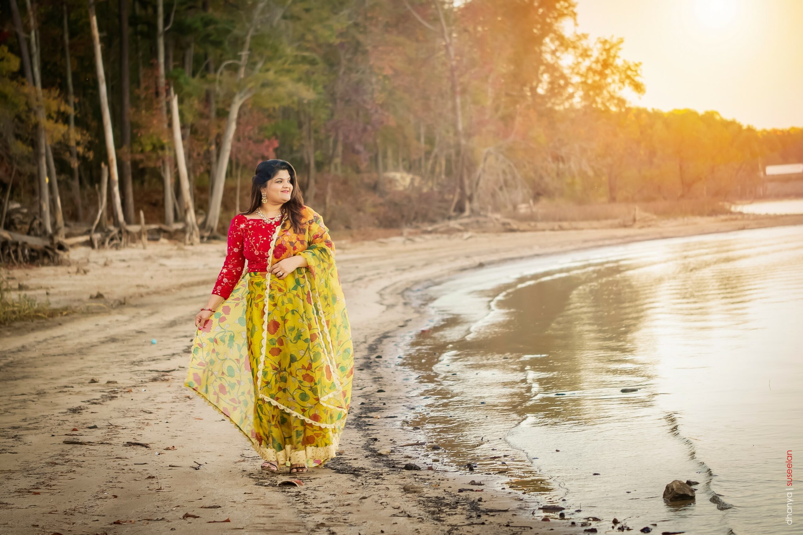 Woman in colorful traditional dress walking along a beach at sunset, trees in background, calm water, warm light.